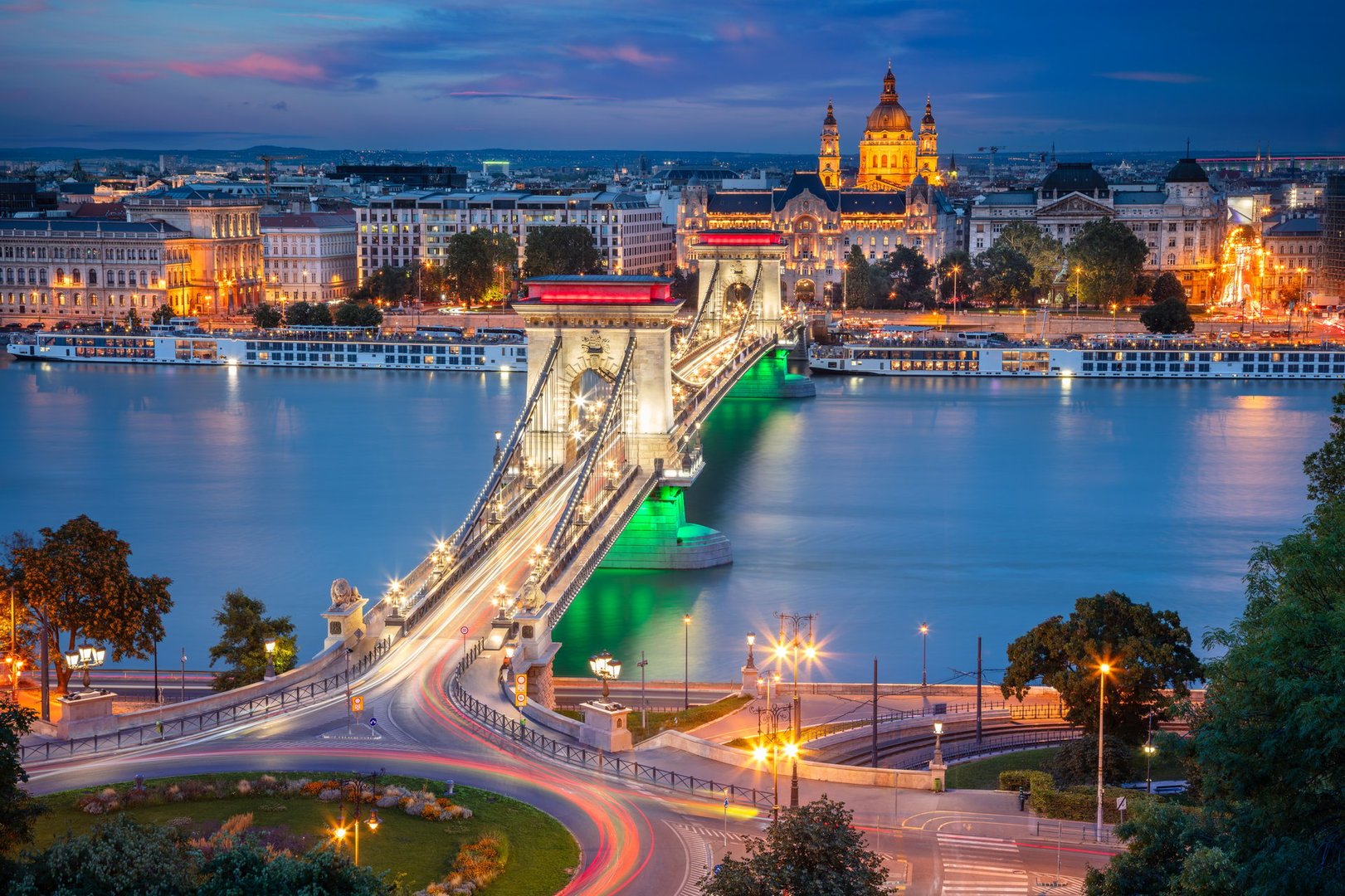 Széchenyi Chain Bridge spans the Danube River in Budapest, Hungary. Illuminated with bright lights, the bridge connects Buda and Pest. A vibrant cityscape forms the backdrop, with St. Stephen's Basilica prominently visible. The swirling light trails from vehicles below indicate evening traffic. The serene blue water reflects city lights, highlighting the architectural elegance of the bridge and surrounding historic buildings, under a deepening dusk sky.