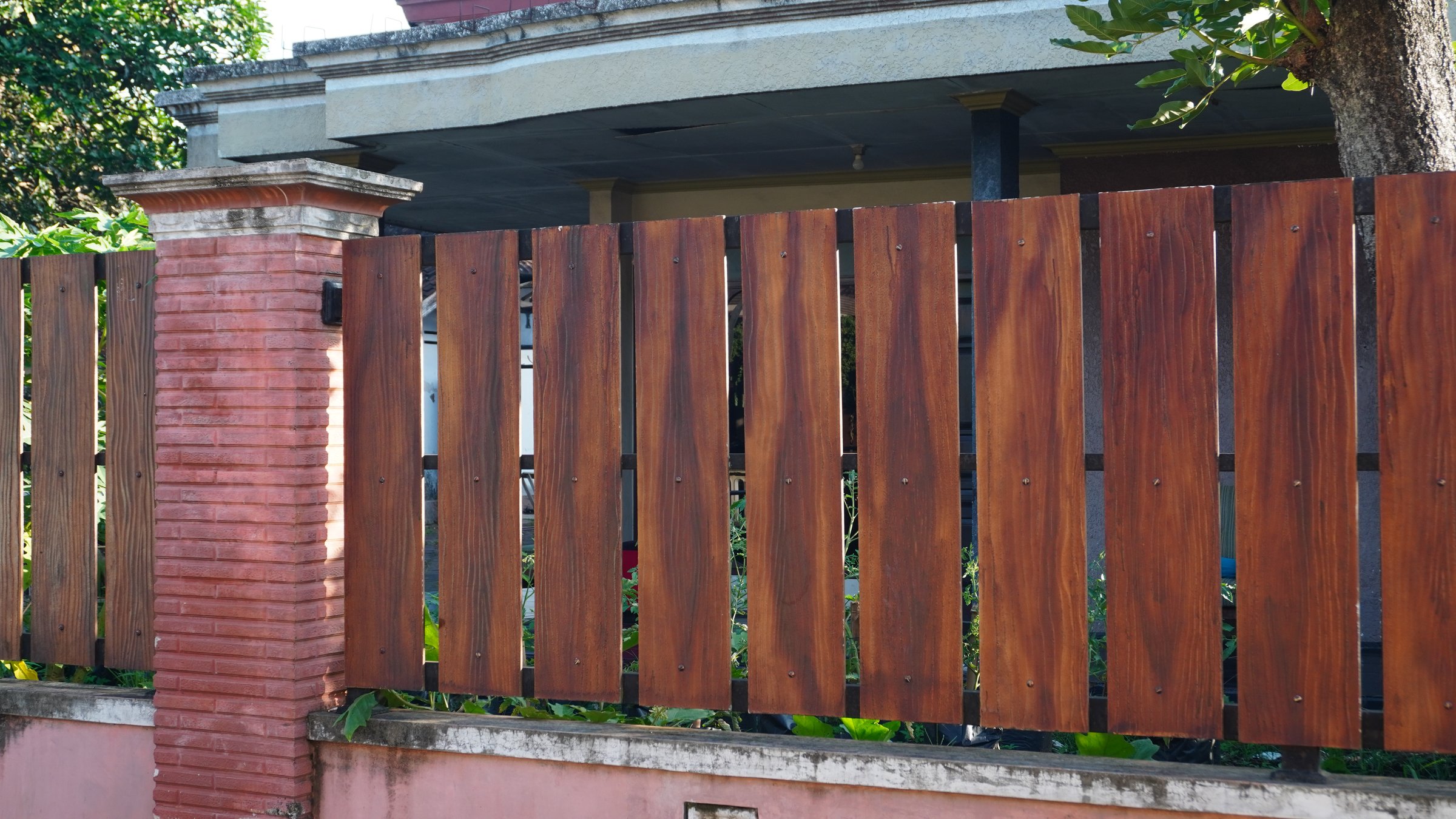 A simple wooden fence stands in a residential yard its brown surface showing natural grain patterns and subtle weathering cracks the boards are arranged with gaps allowing partial views of the surrounding greenery shadows from nearby plants fall across the aged wood.