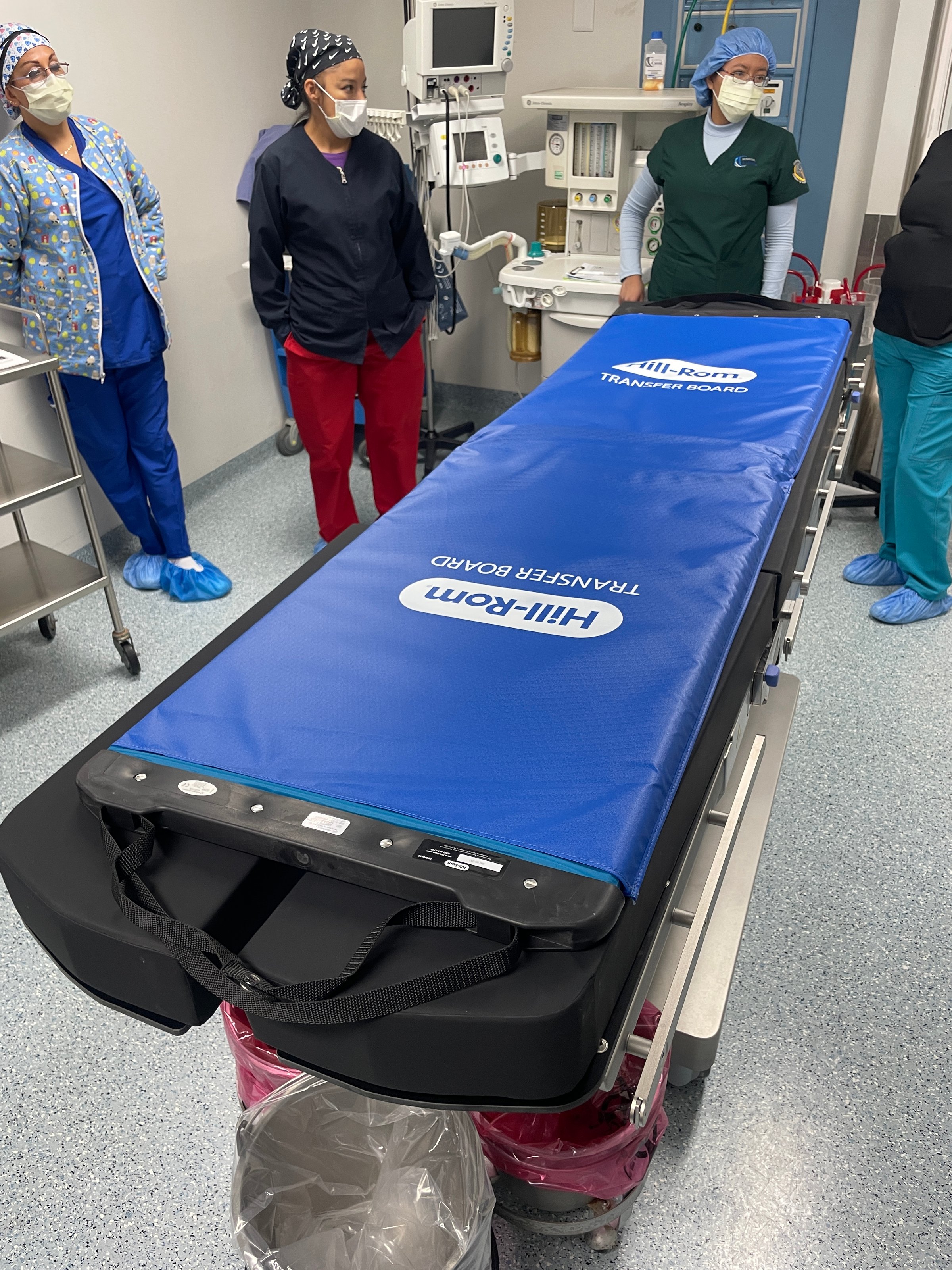 Medical staff standing by a blue patient transfer board in a hospital room, wearing scrubs and masks.