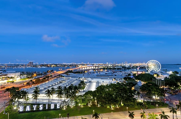 Aerial view of bayside area with illuminated buildings, Ferris wheel, palm trees, and a bridge at dusk.