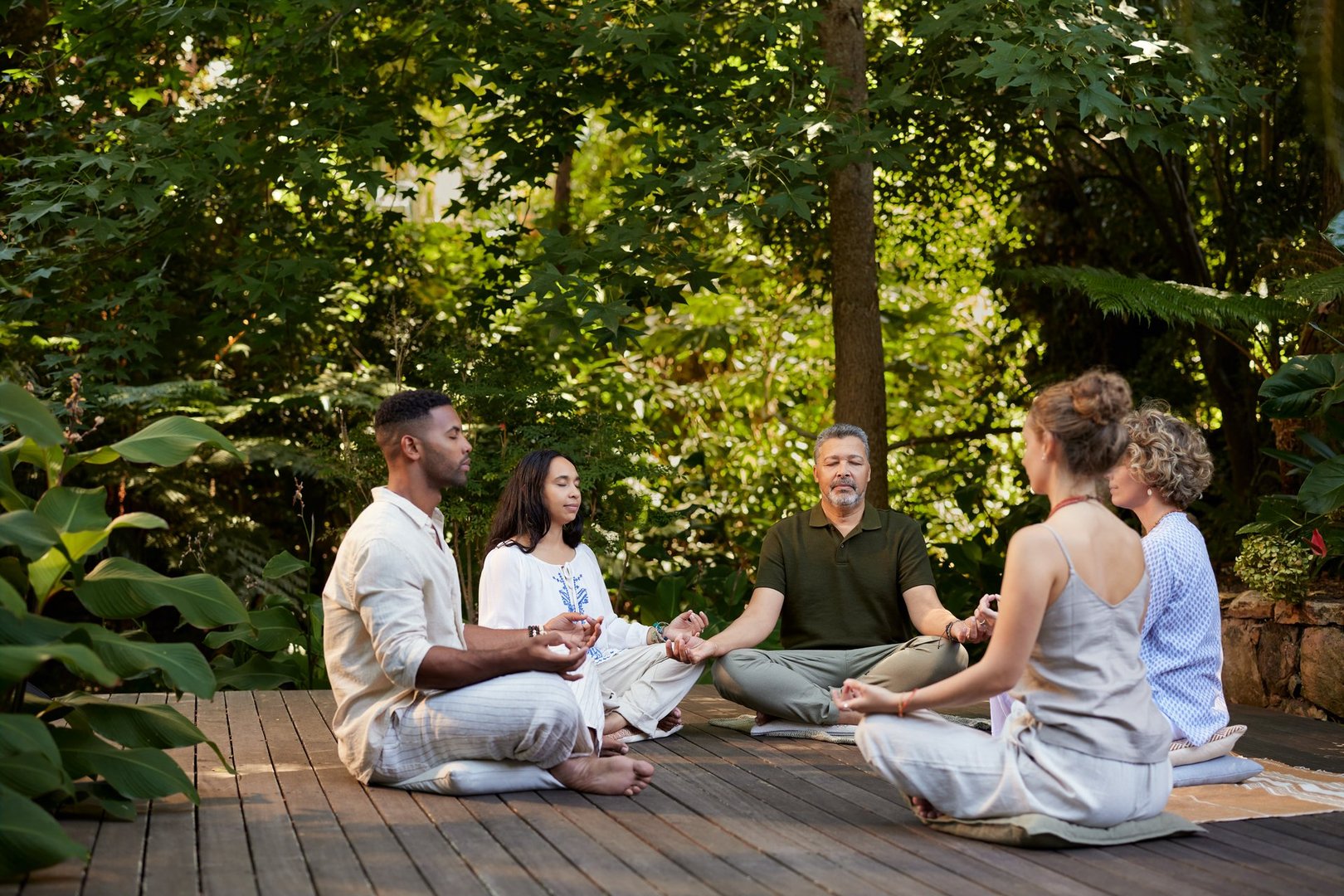 Group of diverse people sitting in a circle and meditating outside in a peaceful green forest. Men and women practicing mindfulness and deep breathing together on a wooden platform in nature. Group of friends sitting in a circle meditating with eyes closed during a restorative yoga session.