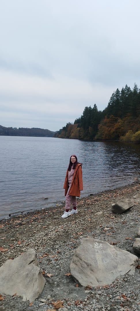 A person in an orange coat stands on a rocky lakeside with trees in the background under a cloudy sky.