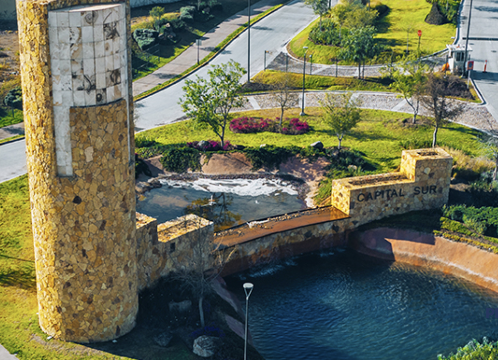 Stone entrance structure with "Capital Sur" sign next to a pond and landscaped greenery, viewed from above.