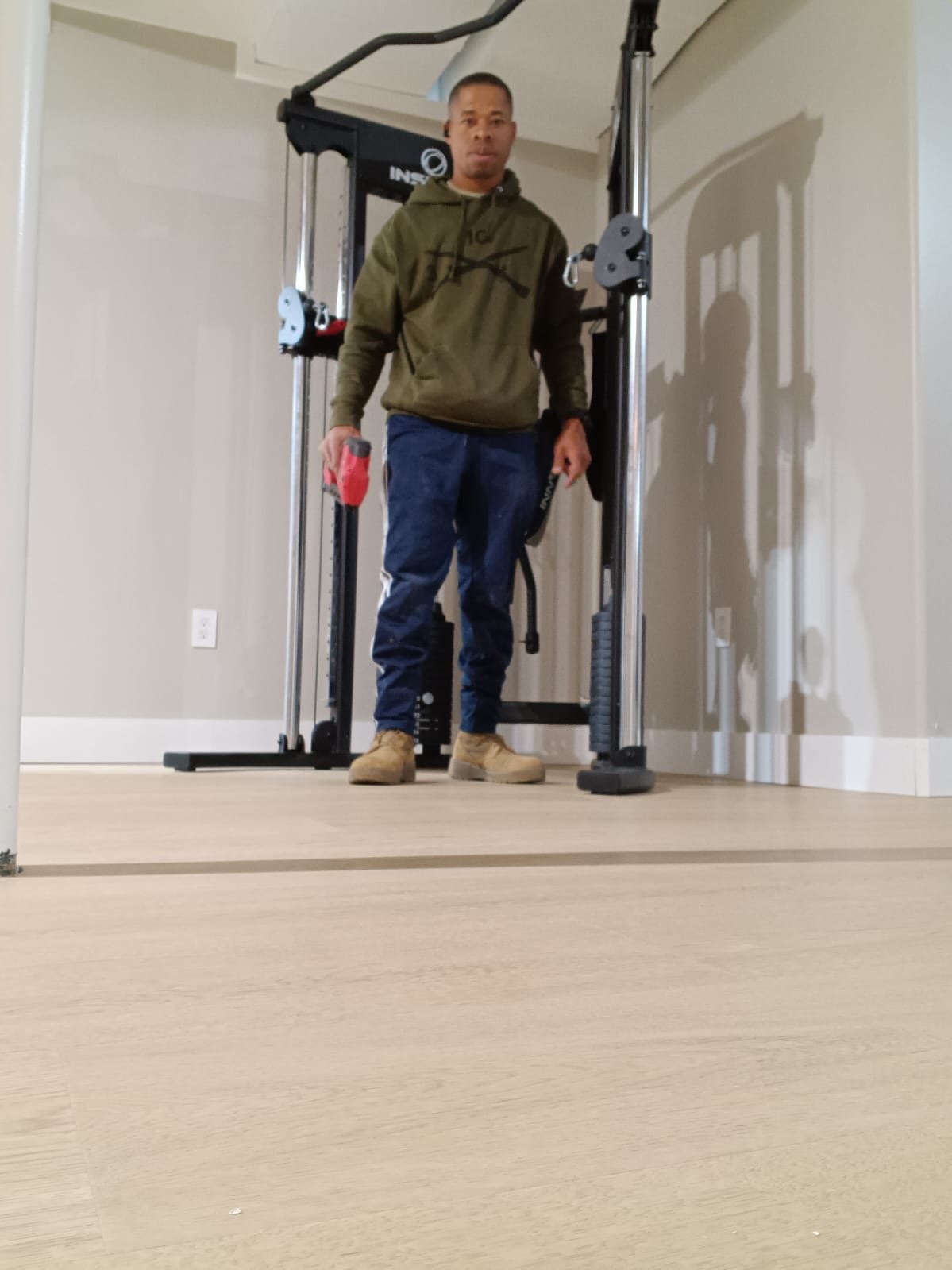 Person standing in a home gym holding a small red object, wearing a green hoodie, blue pants, and tan boots.