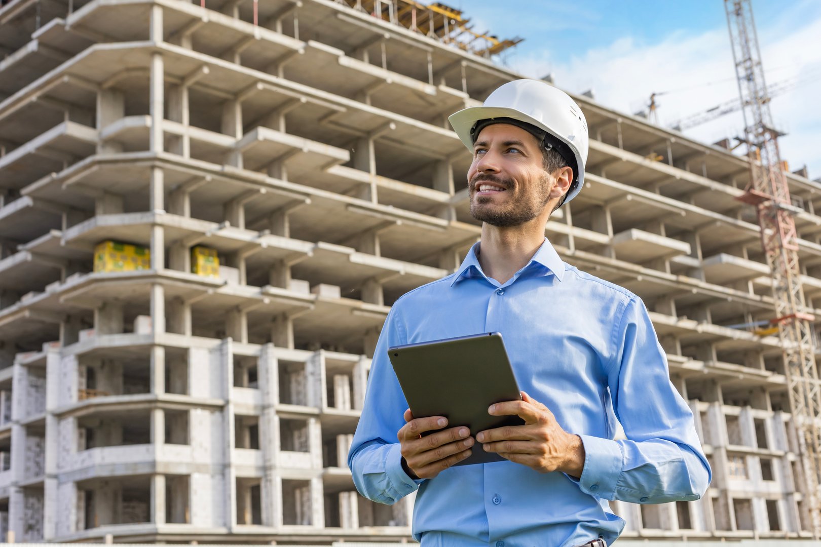 Side view of male engineer standing against background of buildings under construction working in construction helmet.
