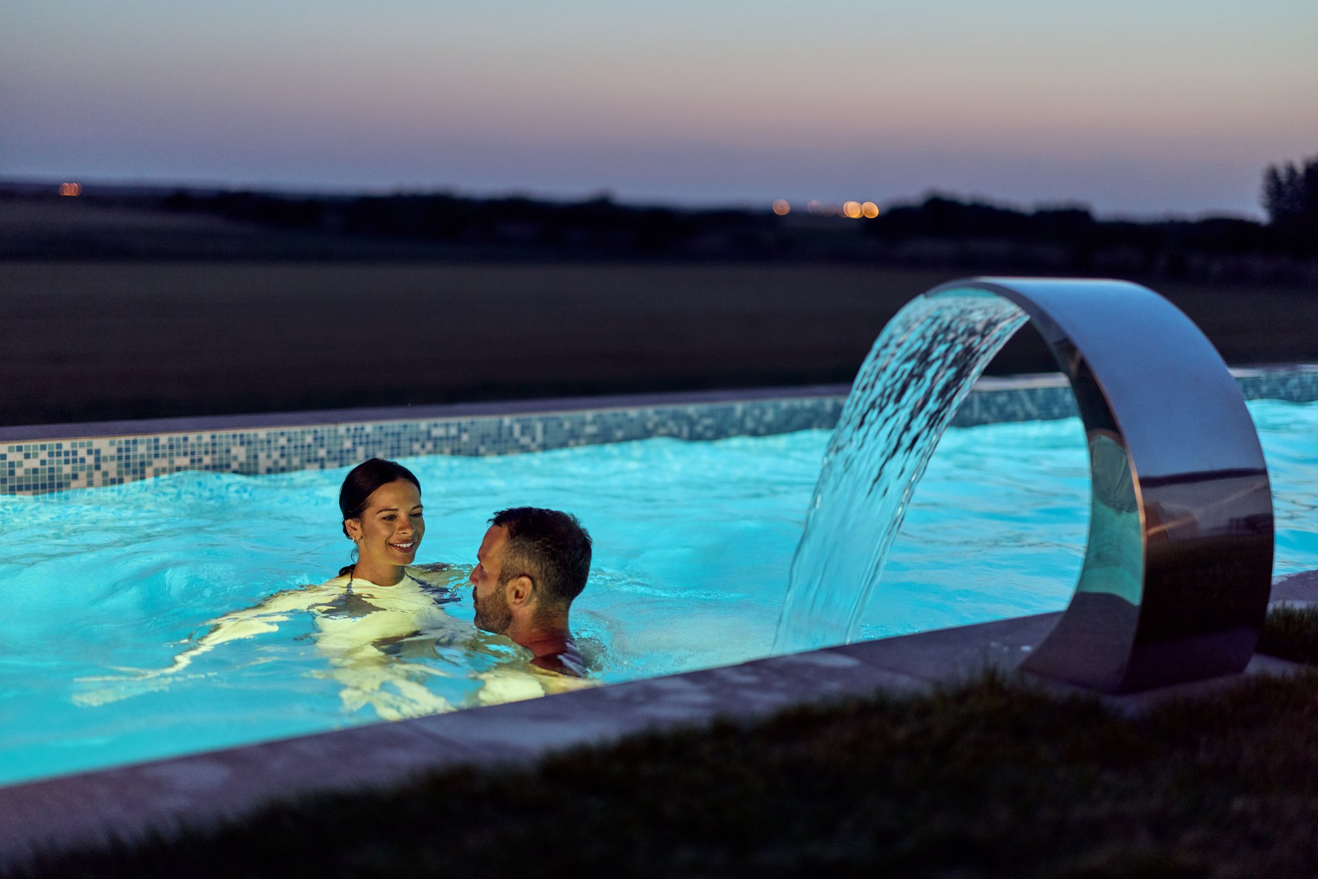 A romantic moment as couple enjoys a glowing pool with cascading waterfall at dusk.