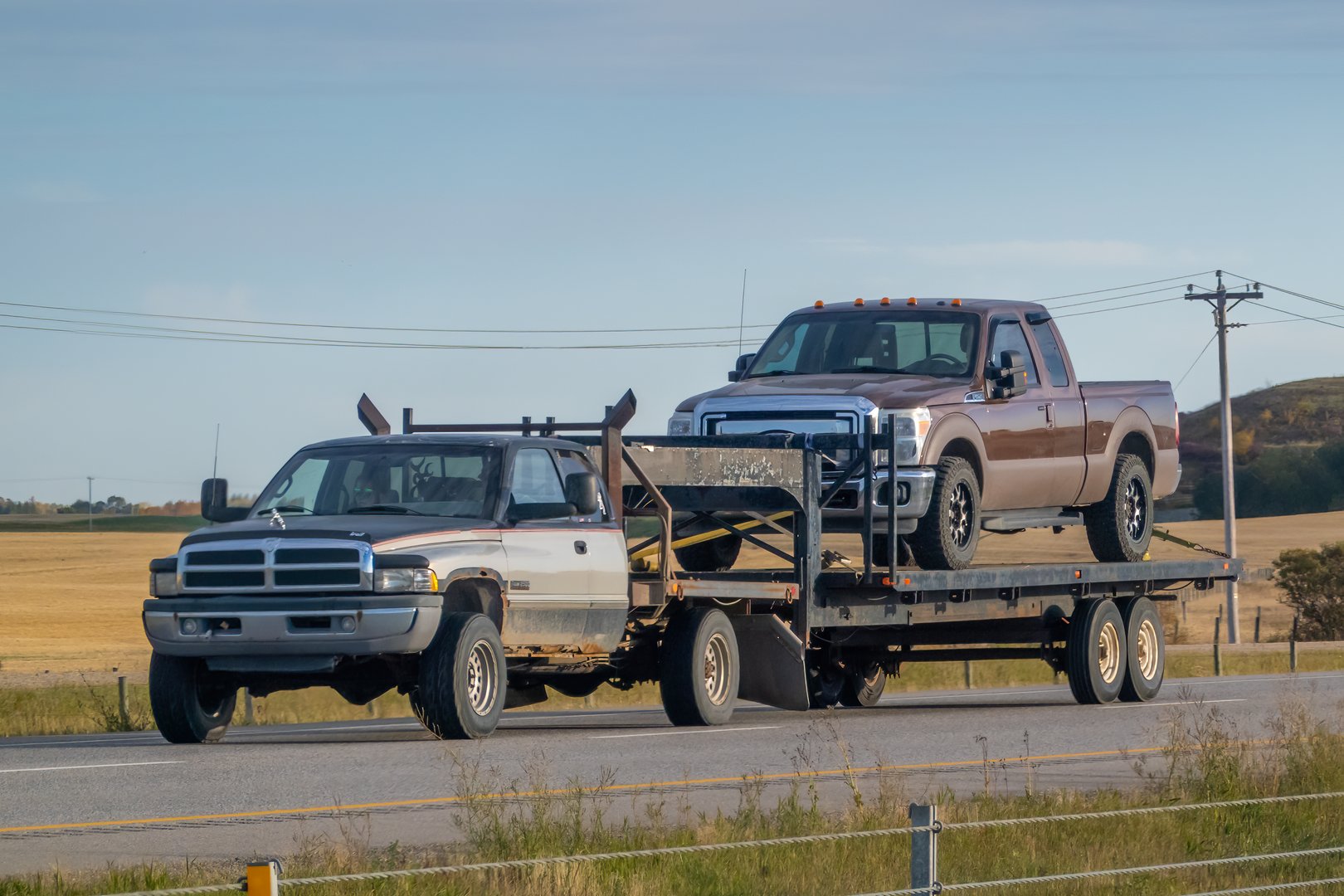 Calgary, Alberta, Canada. Sep 30, 2024. A Dodge Ram pickup truck towing a flatbed trailer with a brown Ford F-Series pickup truck securely fastened on top.