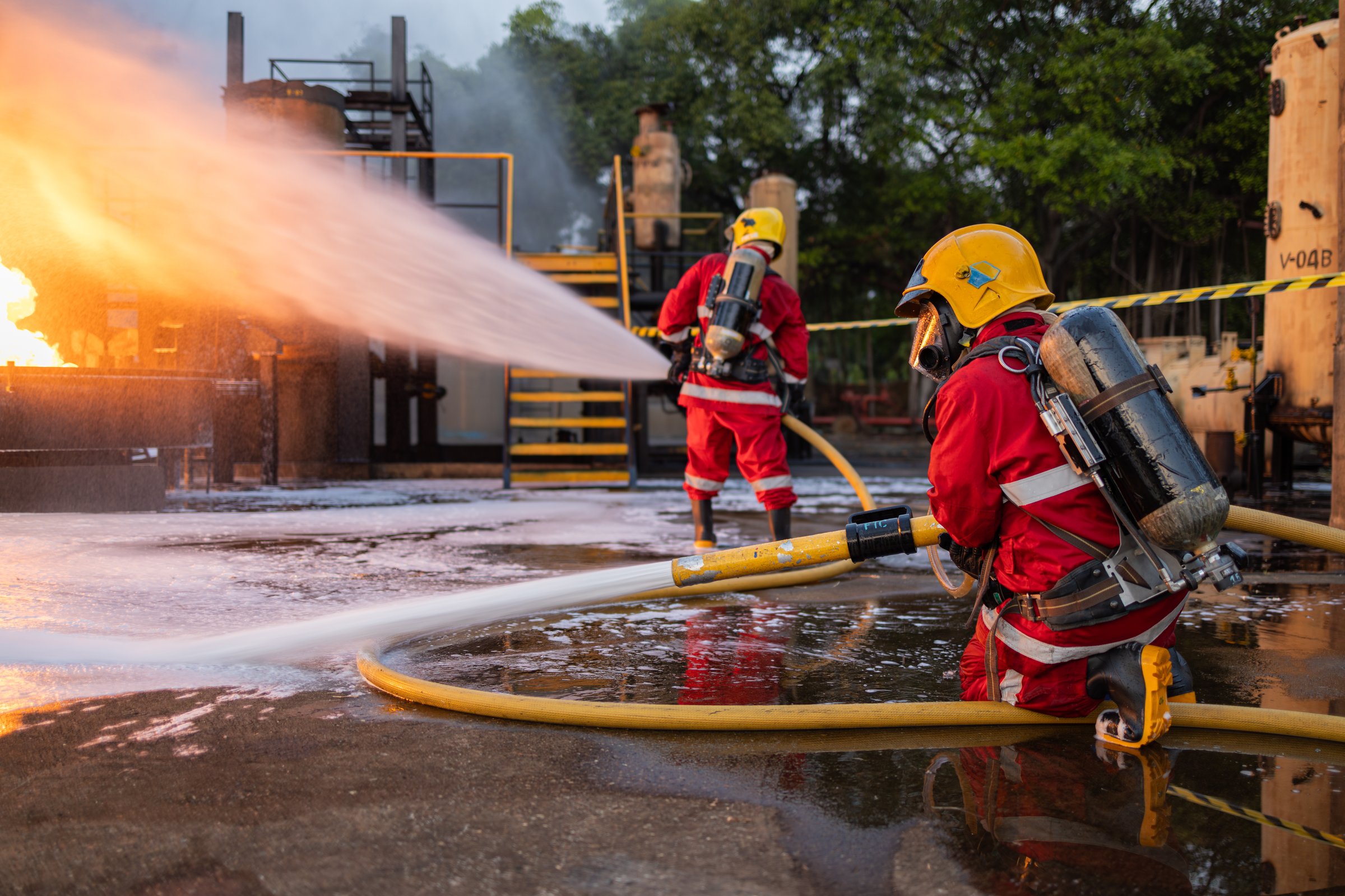 Two firefighters working diligently to suppress roaring flames using foam at an industrial fire scene during dusk.
