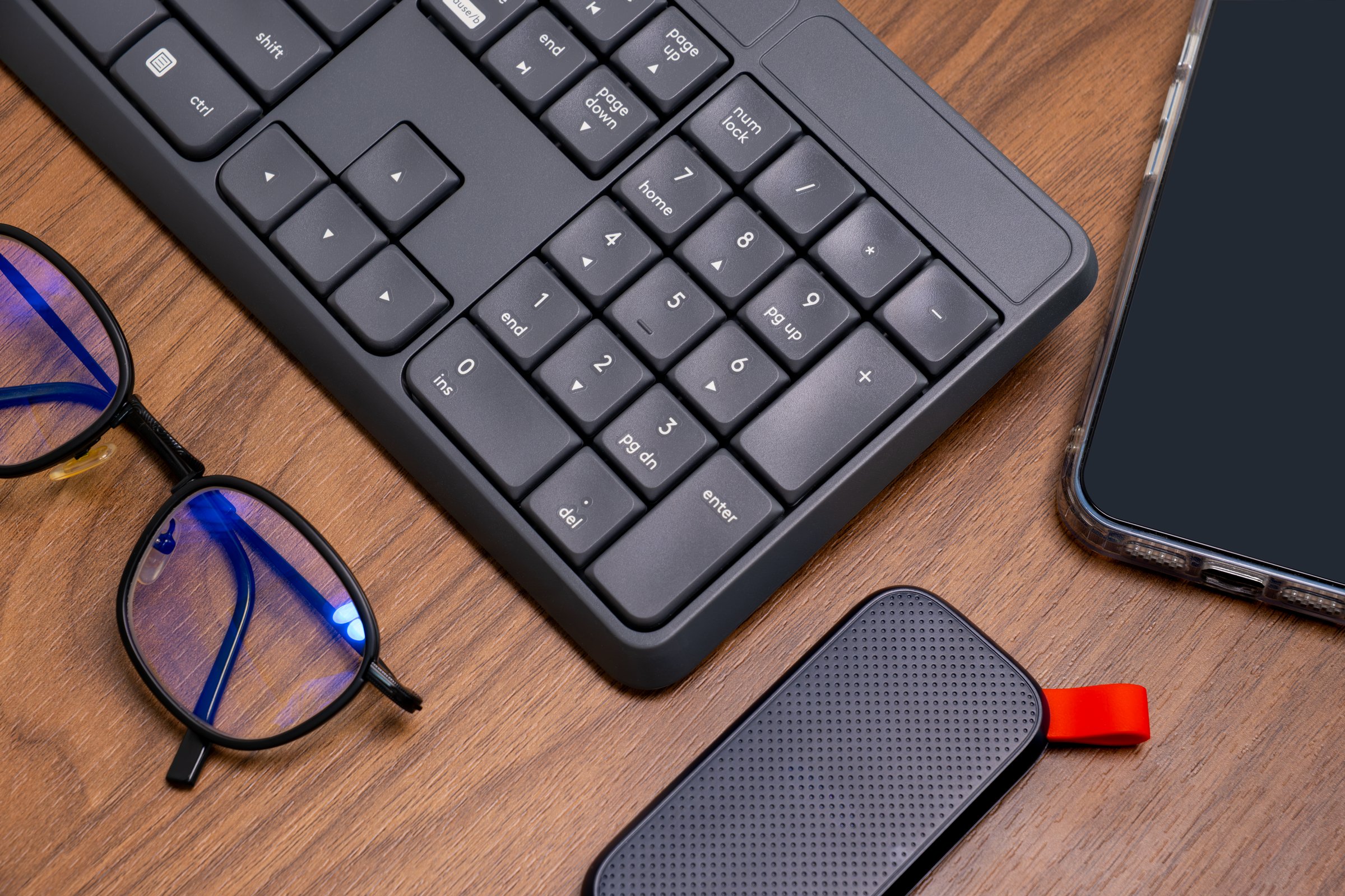 Modern workspace with keyboard, smartphone, hard drive, and blue-lens glasses on wood desk—clean tech setup for digital productivity and device ads.