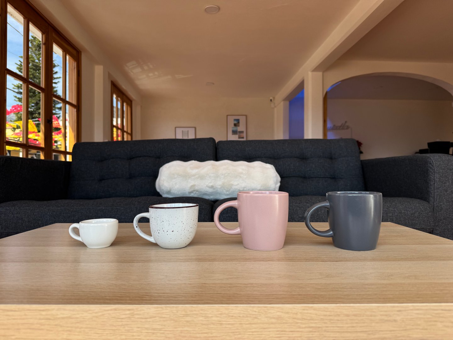 Four mugs of varying sizes and colors on a wooden table in a living room with a sofa and large windows in the background.