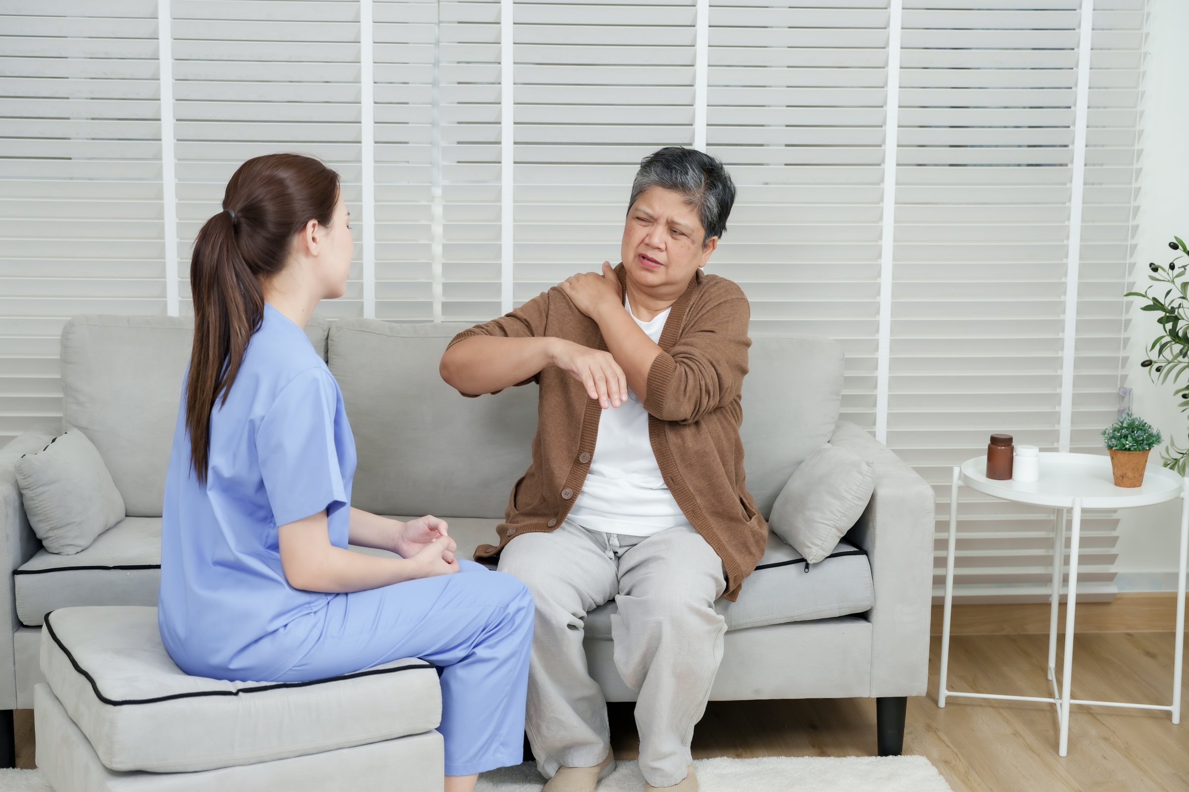 Senior asian woman seated on sofa raising arm while pointing at painful shoulder as female physiotherapist observes closely during orthopedic consultation in home-like rehabilitation room