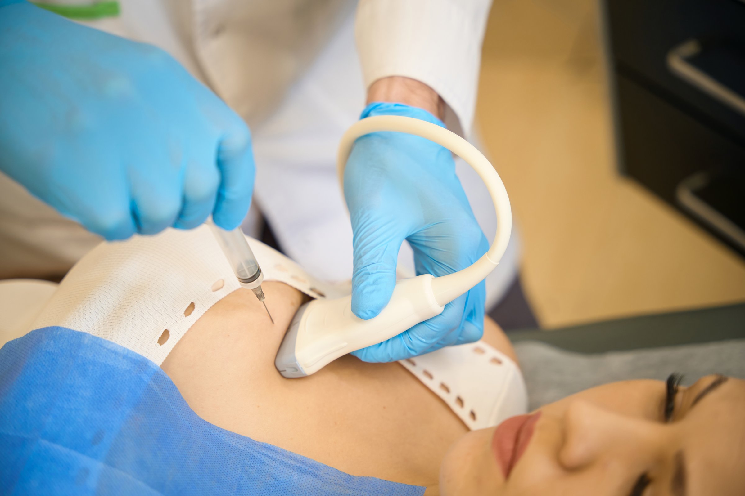 Doctor in nitrile gloves inserting syringe needle into female patient breast next to ultrasound probe