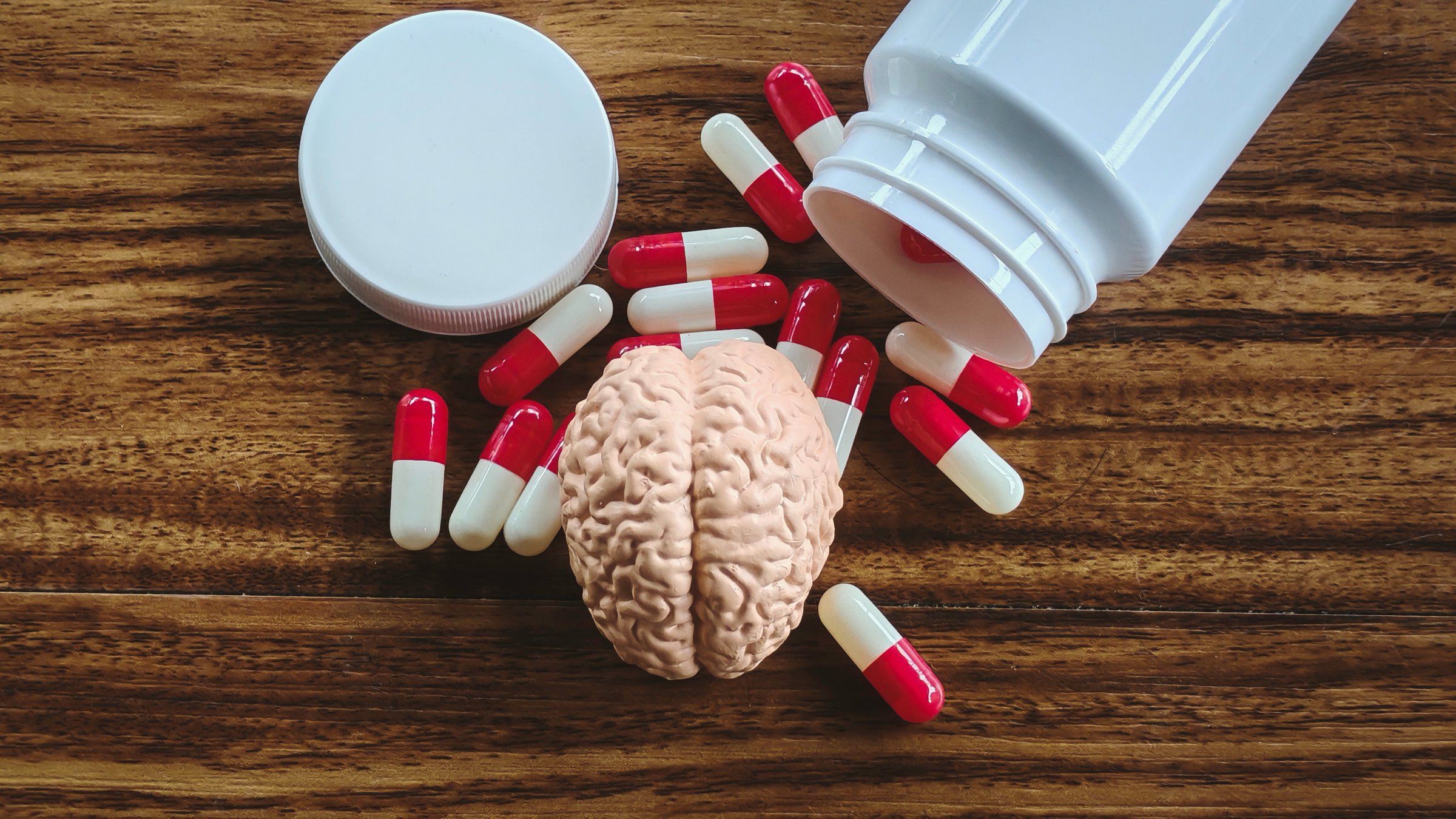 Miniature Brain Model and Spilled Red and White Pills from a Bottle on a Wooden Surface, Representing Mental Health, Neurological Disorders, and Psychiatry Treatment.