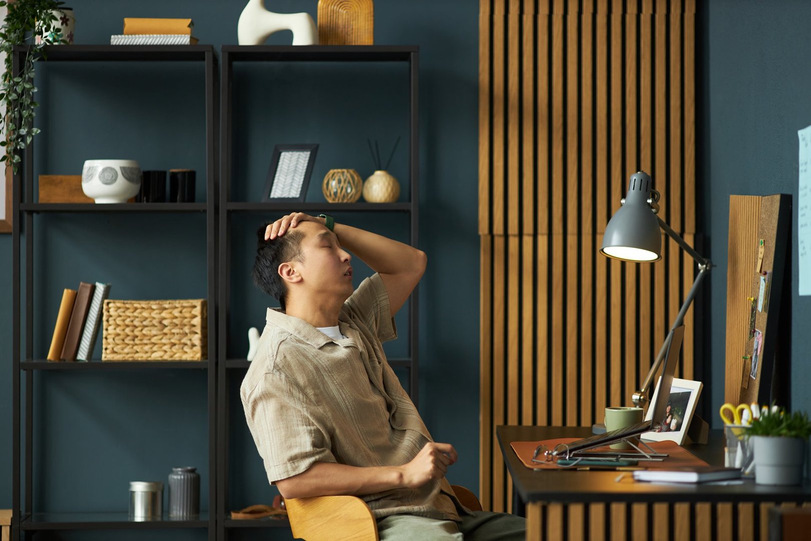 Asian young adult man sitting at desk, holding head with hand appearing stressed while looking at computer monitor in modern home office workspace with shelves in background