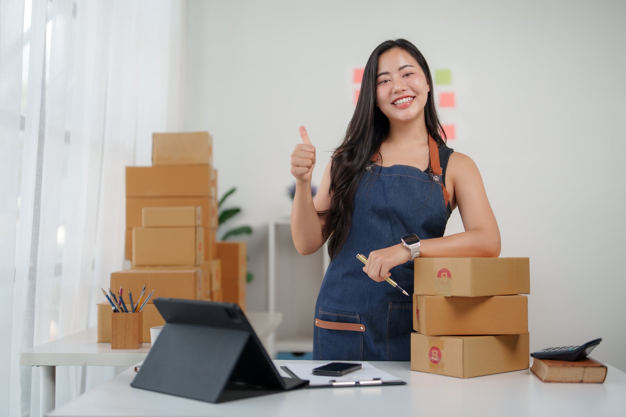 Young asian woman entrepreneur giving a thumbs up while working confidently in a home office, showcasing success and motivation
