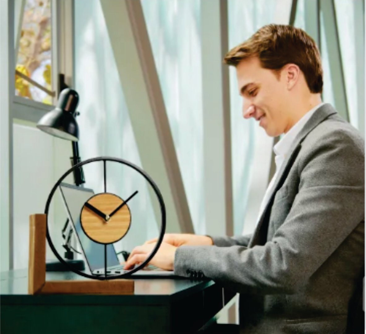 Man in a gray blazer working on a laptop at a desk with a modern circular clock, under a desk lamp near large windows.