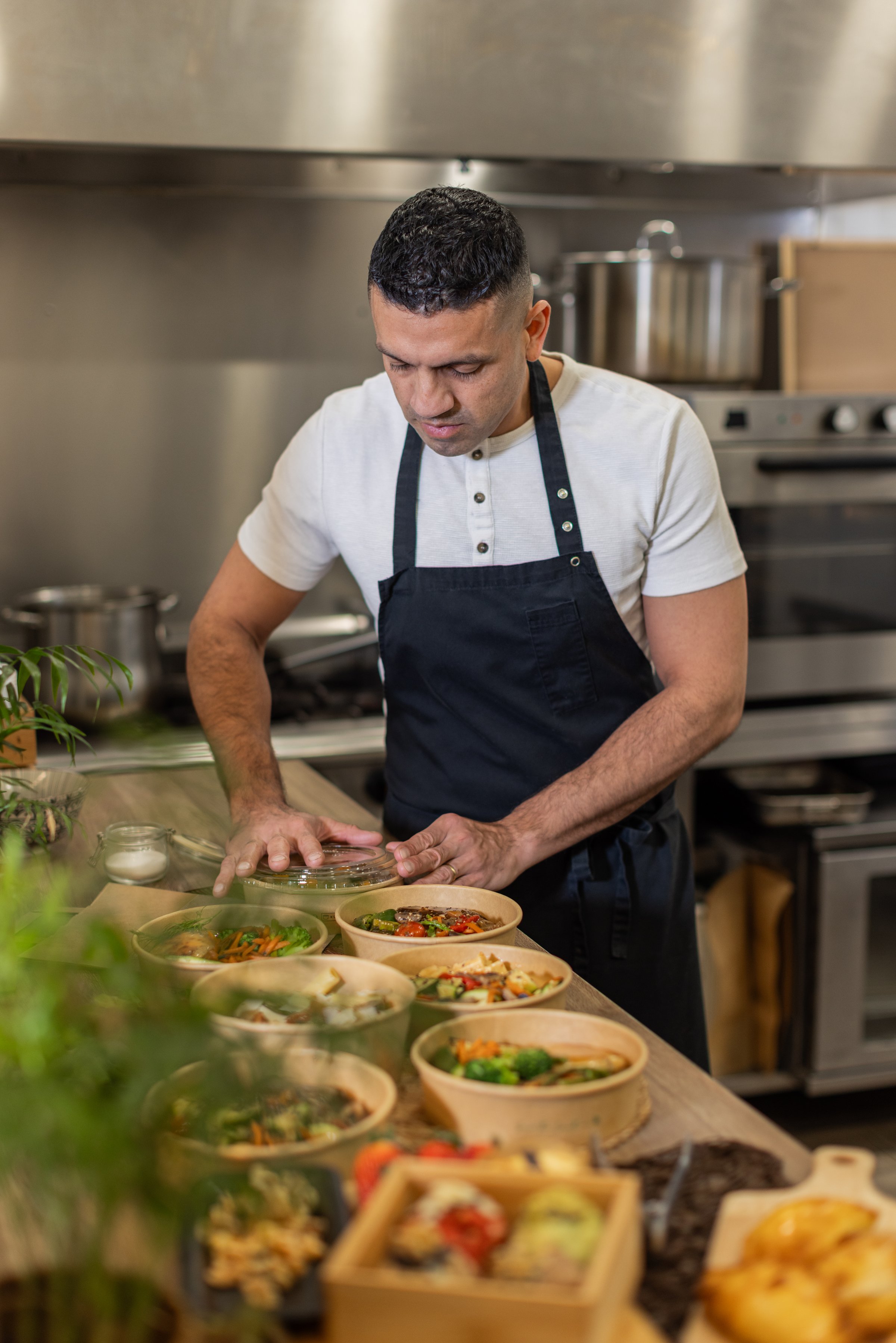 A chef in a black apron carefully prepares and packs takeaway meals in eco-friendly bowls. The setting is a professional kitchen with stainless steel appliances, fresh ingredients. Vertical copy-space