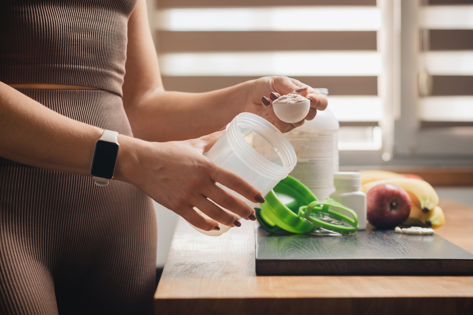 Athletic woman in sportswear with measuring spoon in her hand puts portion of whey protein powder into a shaker on wooden table with amino acid white capsules, bananas and apple, making protein drink.