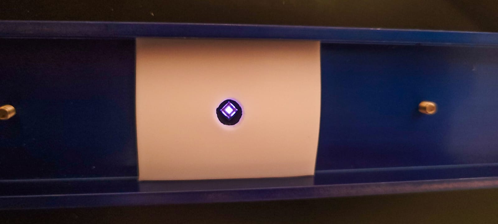 A blue and white device with a small glowing blue light in the center, mounted on a wall with two gold screw heads visible.