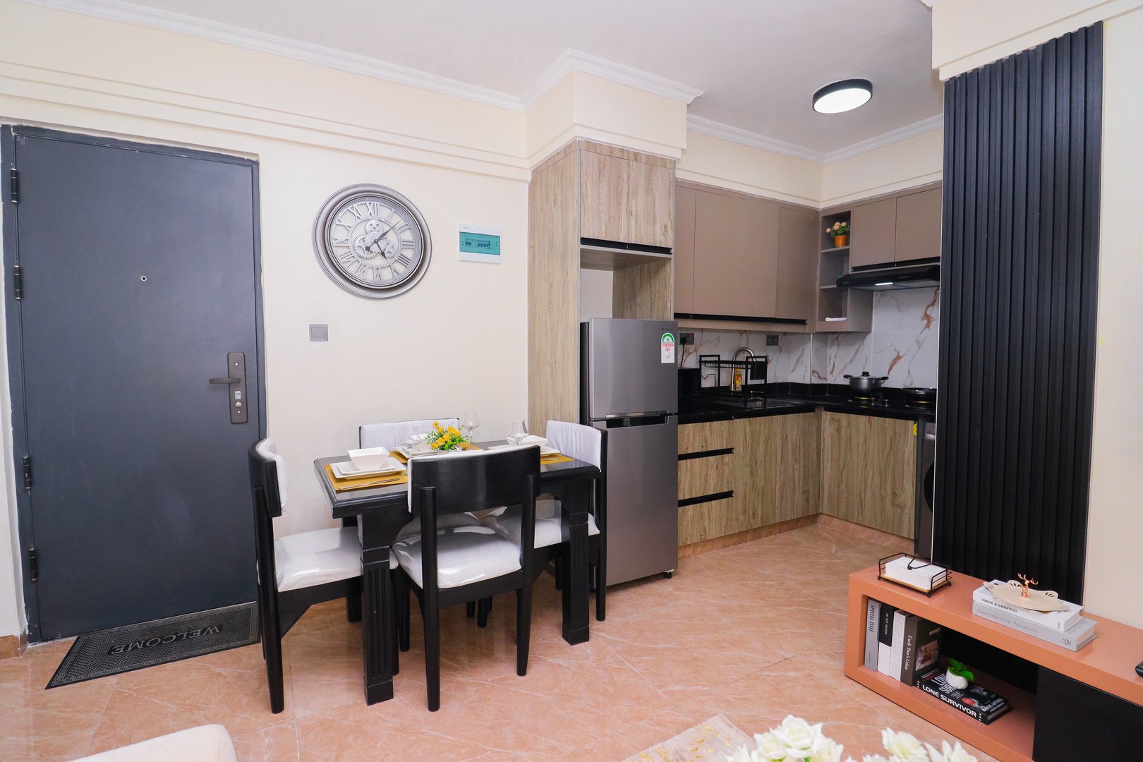 Modern kitchen with dining area, refrigerator, stove, wall clock, and gray door. Minimalist decor with neutral colors.