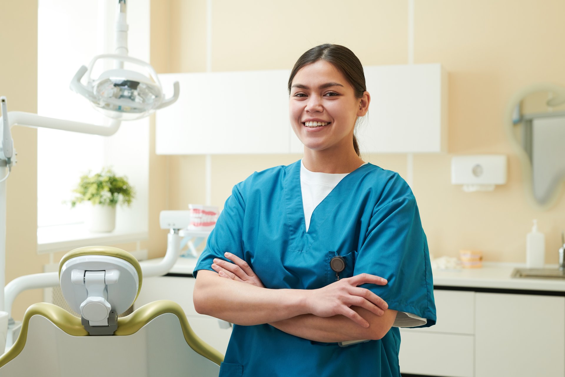 Portrait of young adult woman dental professional standing in dental clinic smiling with arms crossed, dental chair and equipment visible in background
