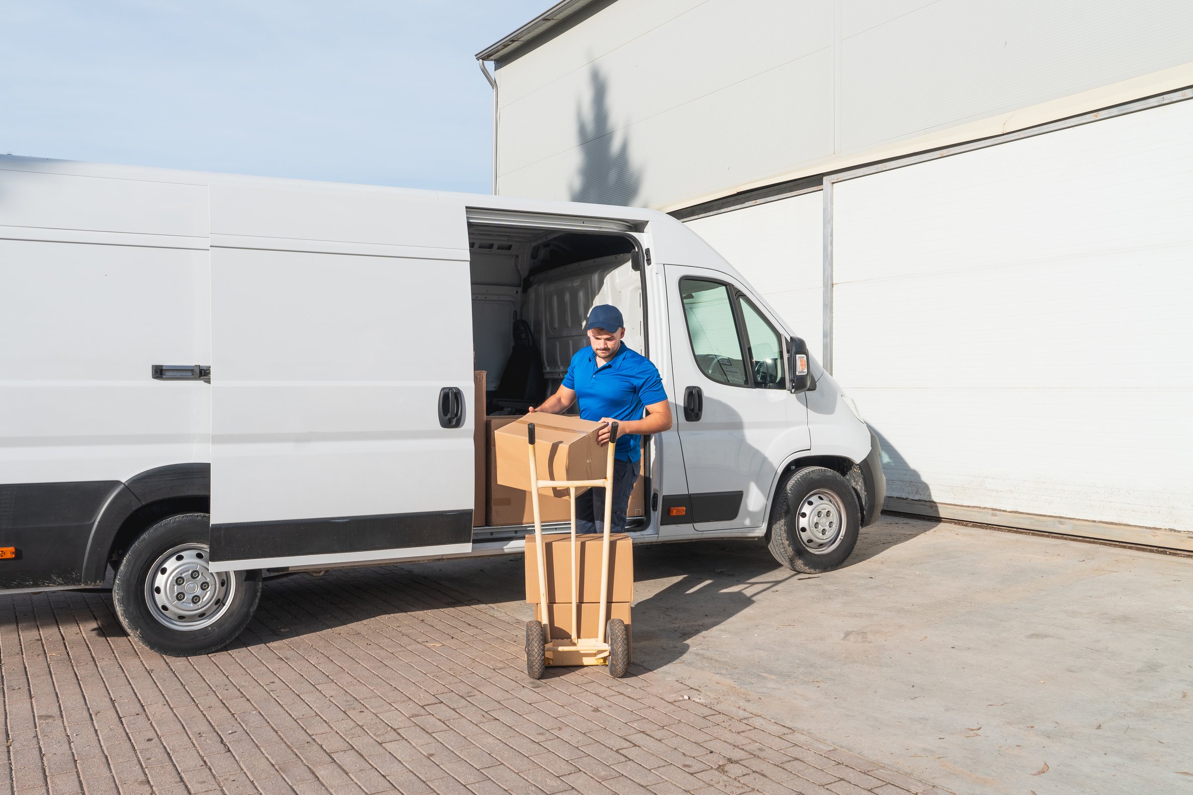 Courier in uniform unloading cardboard boxes from a white van using a hand truck outside a warehouse - Delivery Concept