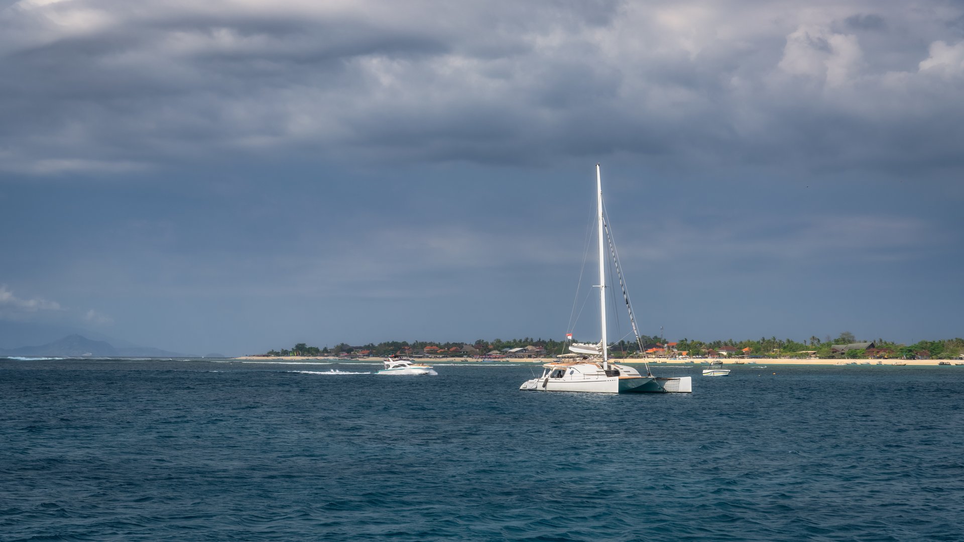 A beautifully tranquil scene showcasing a sailboat gliding gracefully on calm waters, positioned near picturesque coastline that is contrasted by dramatic skies above, Nusa Lembongan, Bali, Indonesia