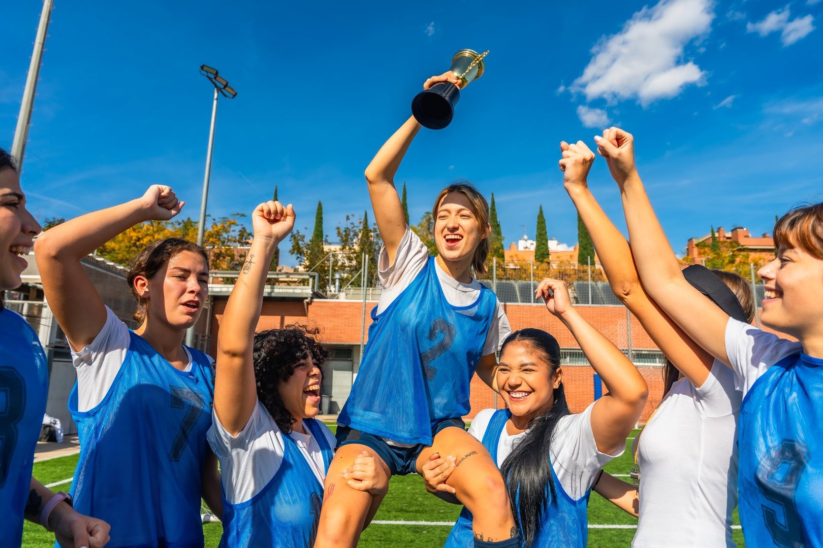 Women's soccer team members smiling, cheering, and lifting a championship trophy on the field after winning a match, celebrating their success and teamwork