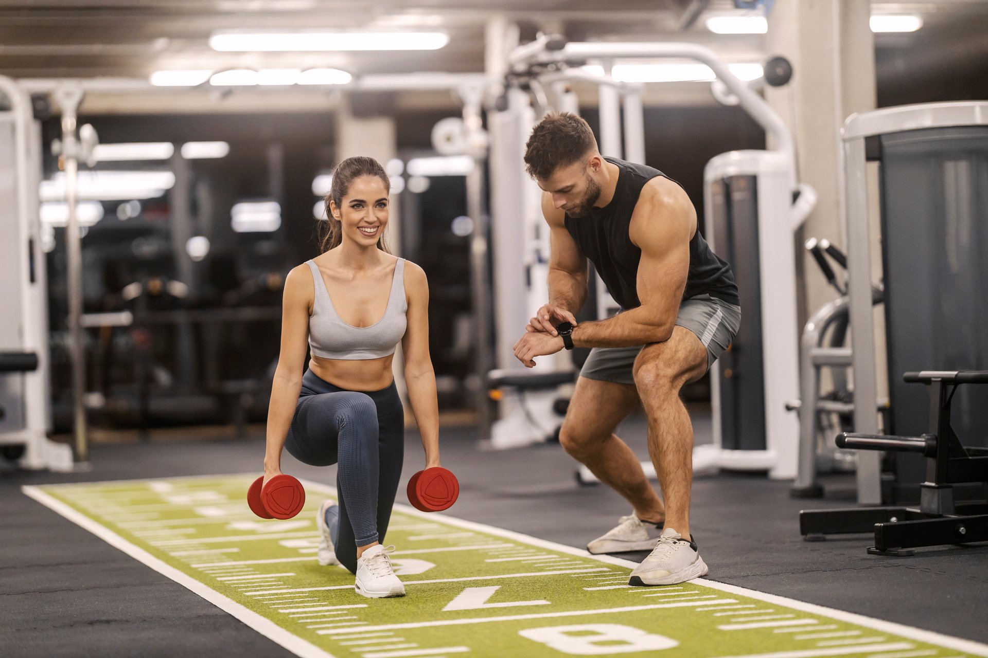 Smiling muscular sportswoman doing lunges with dumbbells in hands with her coach.