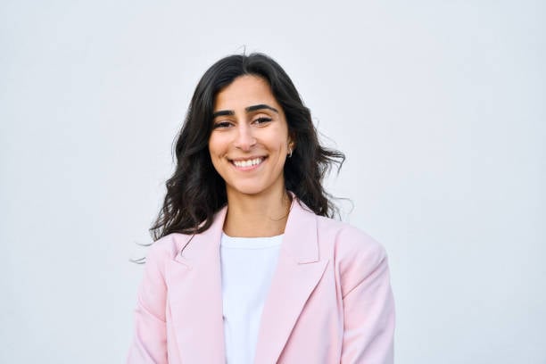 Smiling woman with long dark hair wearing a pink blazer and white shirt against a plain background.