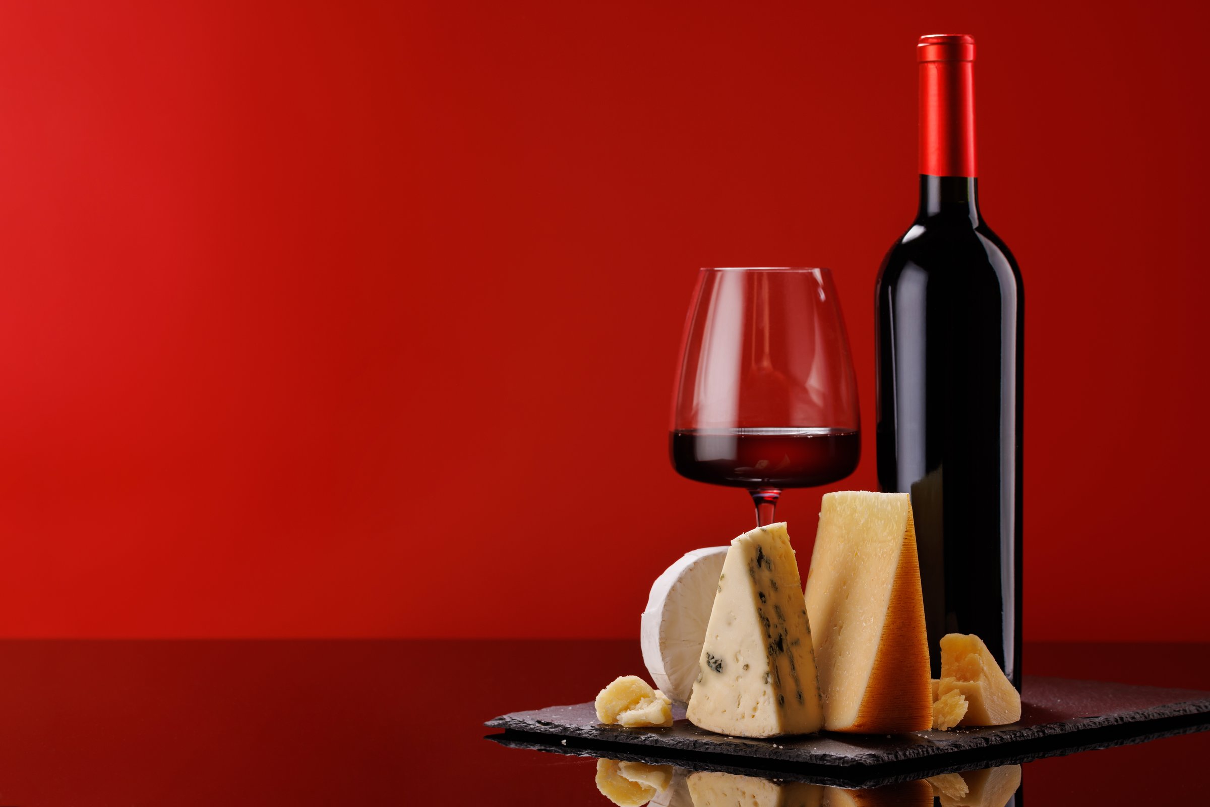 A refined selection of aged cheeses presented on a board, accompanied by a glass of wine, set against a red background. A perfect setting for a gourmet indulgence