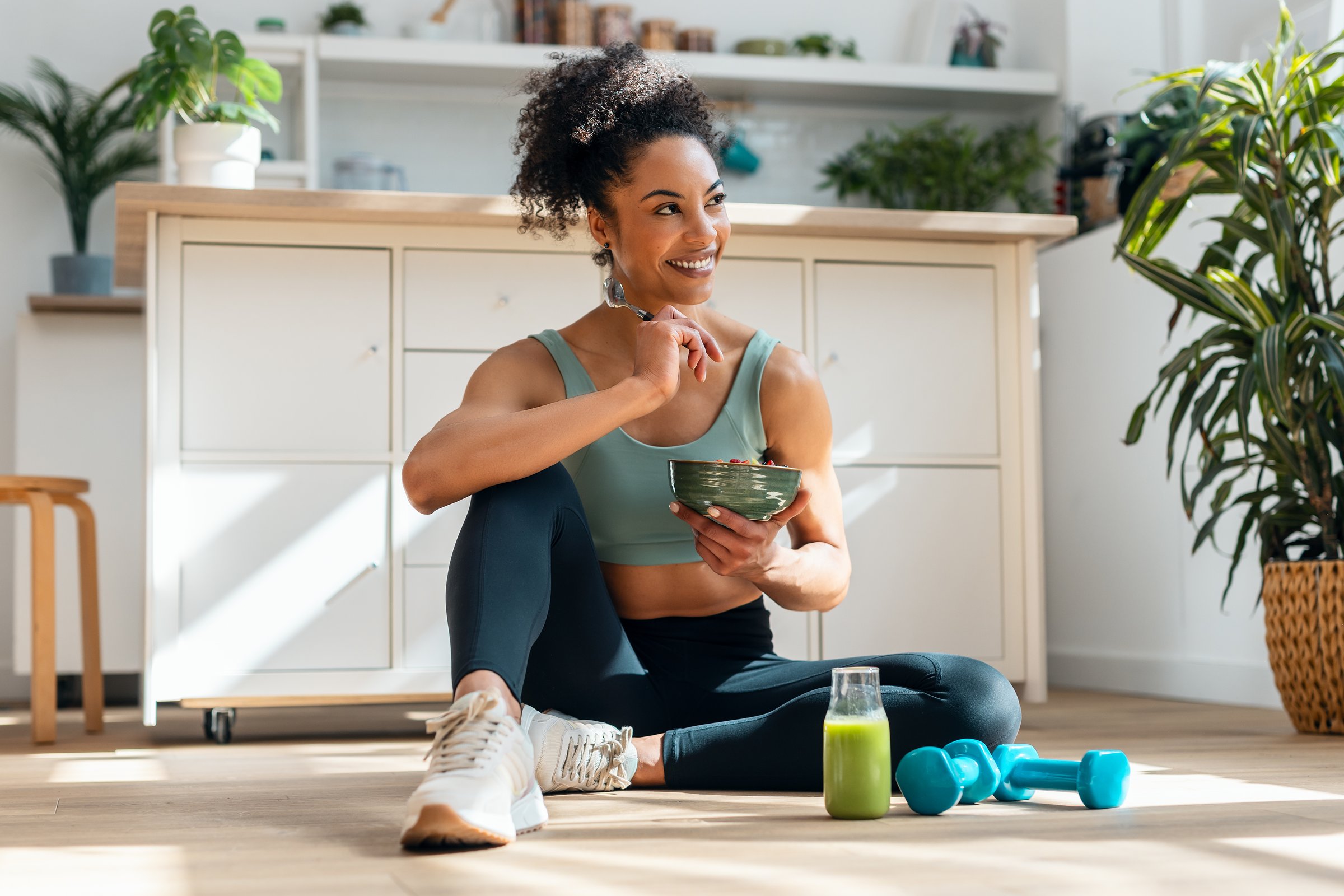 Shot of athletic woman eating a healthy fruit bowl while sitting on floor in the kitchen at home