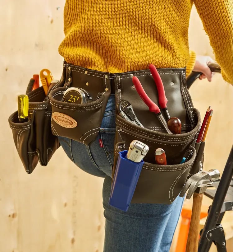 Person wearing a tool belt with various tools including pliers, measuring tape, and hammer, standing in a workshop.