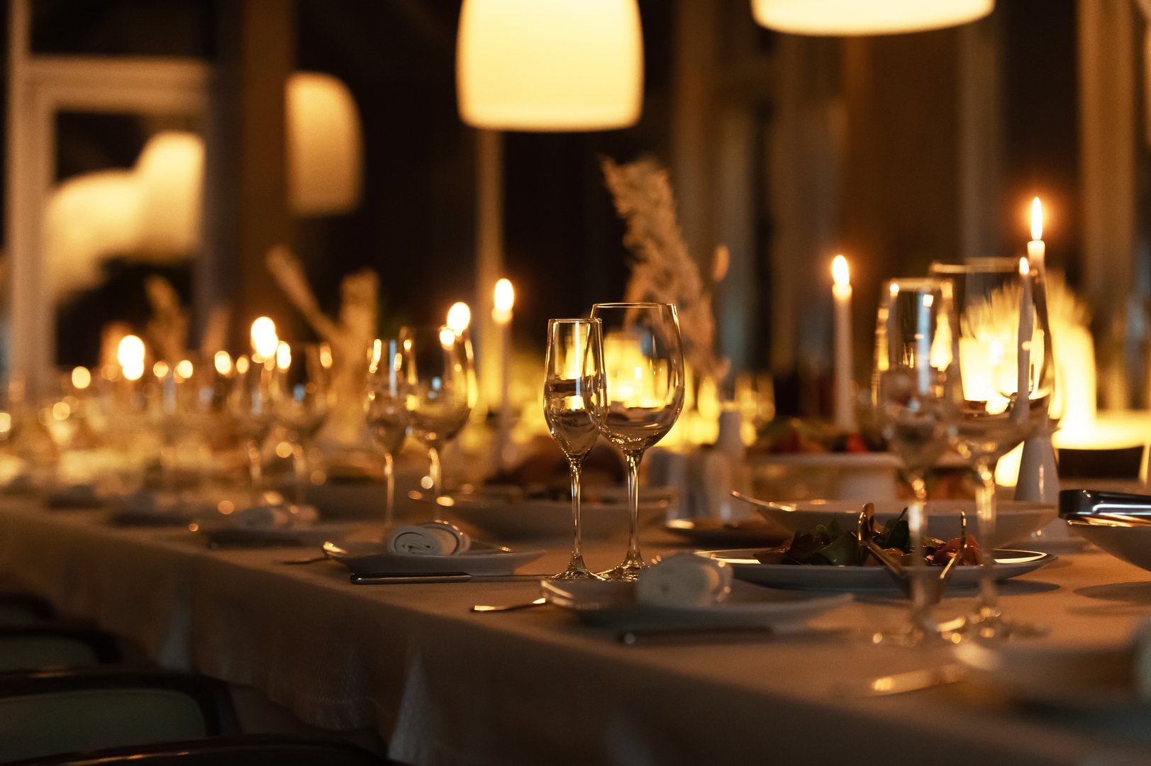 Set in a modern restaurant, a long table is beautifully arranged with glassware and glowing candles for dinner.