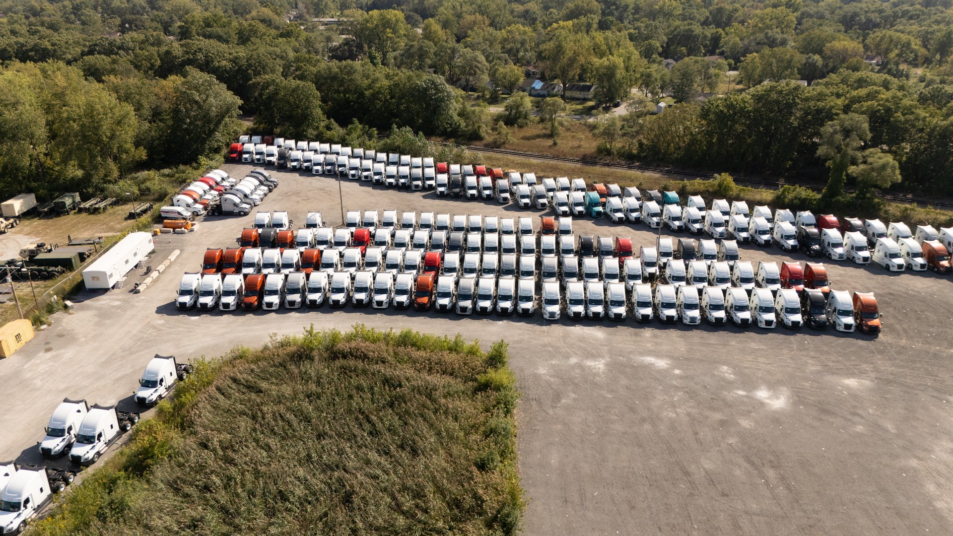 Large lot of parked trucks, various colors, orderly rows, with distant buildings in background under cloudy sky.