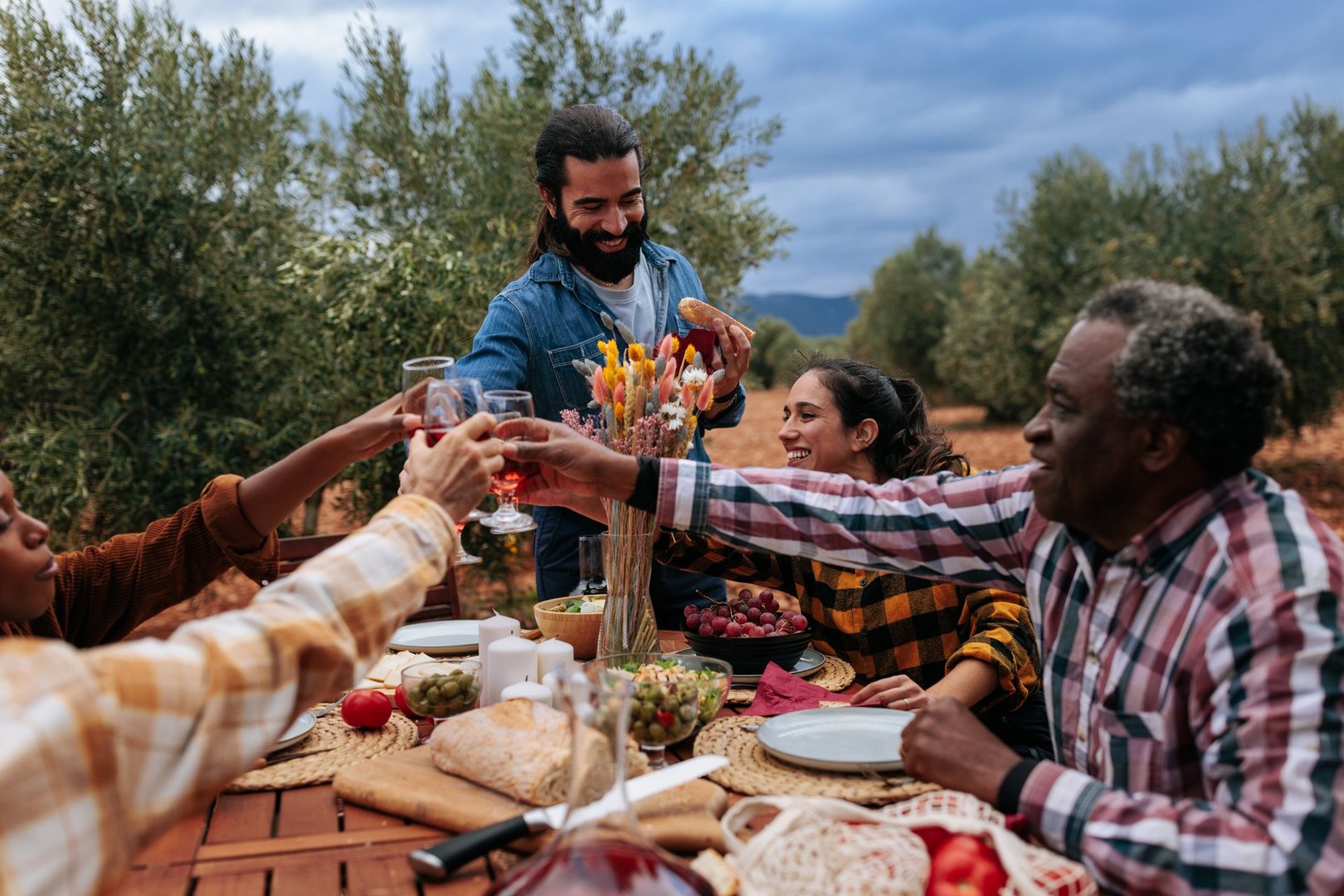 Group of multiracial farmers enjoying a meal and toasting with wine at a table set outdoors in an olive grove