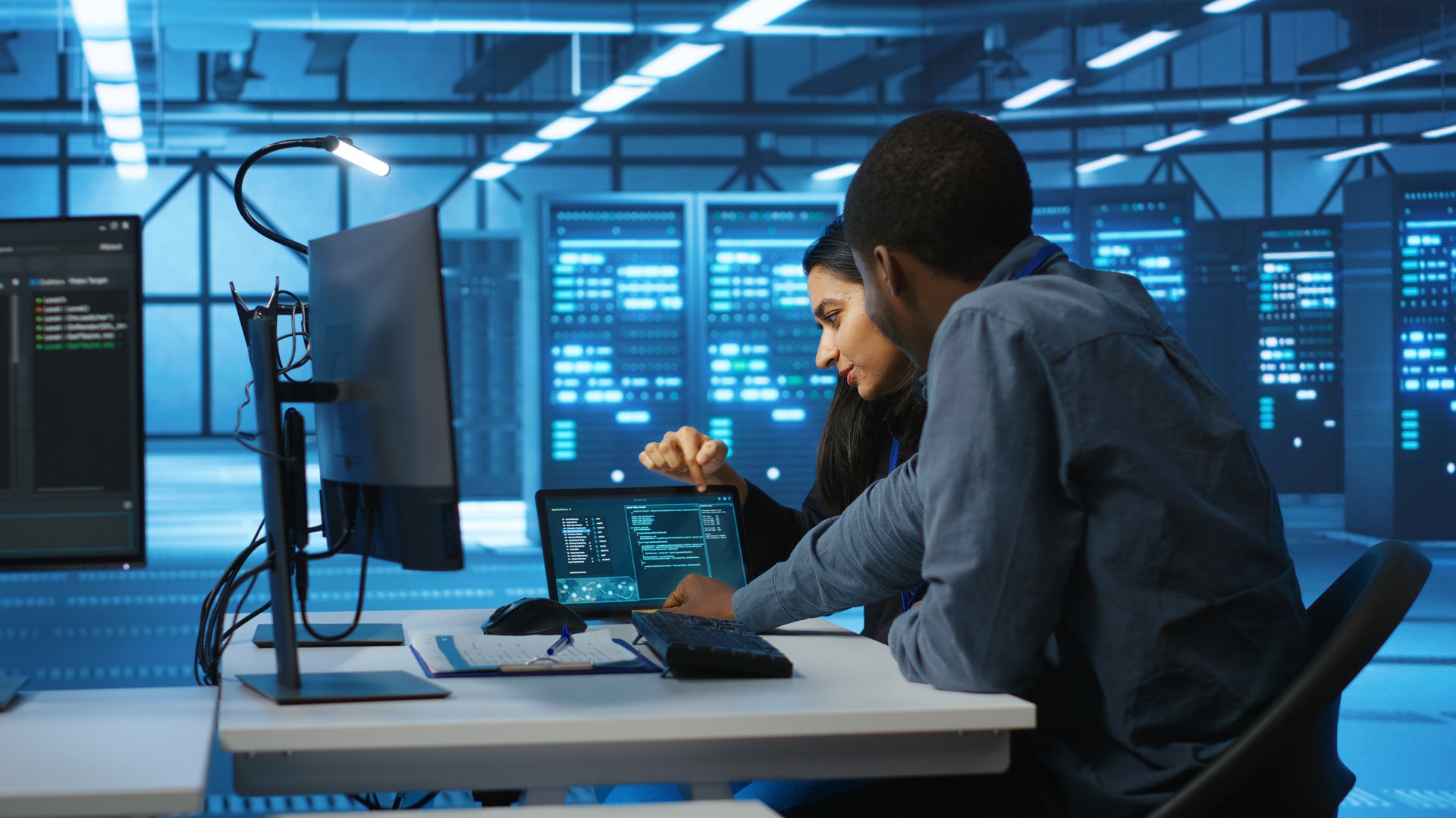 Multiethnic coworkers doing system analysis of server farm supercomputers using tablet. Diverse team of engineers looking at parameters in data center housing storage infrastructure