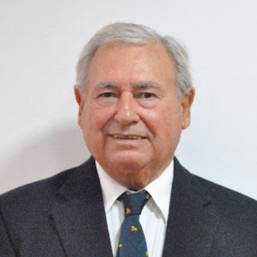 Elderly man with gray hair wearing a dark suit and patterned tie, standing against a plain white background.