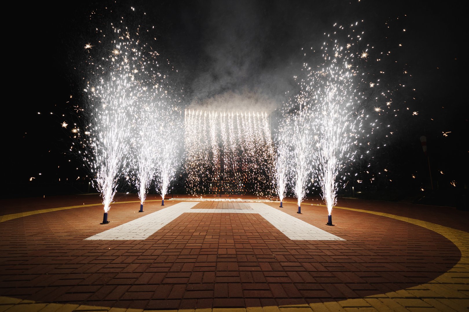 A vibrant celebration takes place at night, featuring dazzling fireworks and fountain-like spark effects illuminating the area, creating a festive atmosphere for attendees.