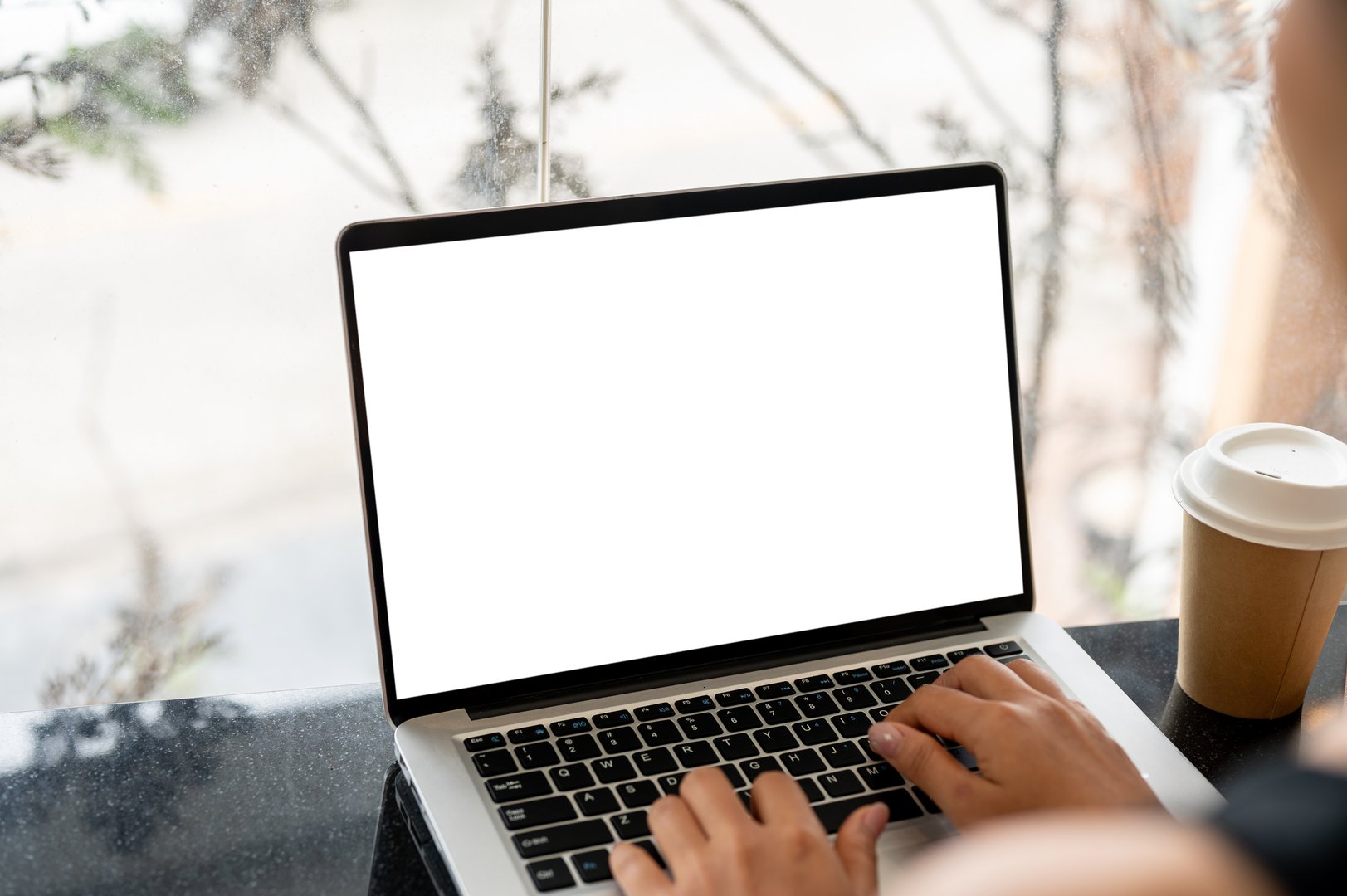 A close-up image of a woman working on her laptop computer at a table in a coffee shop, typing on the laptop keyboard. the laptop with a white screen mockup
