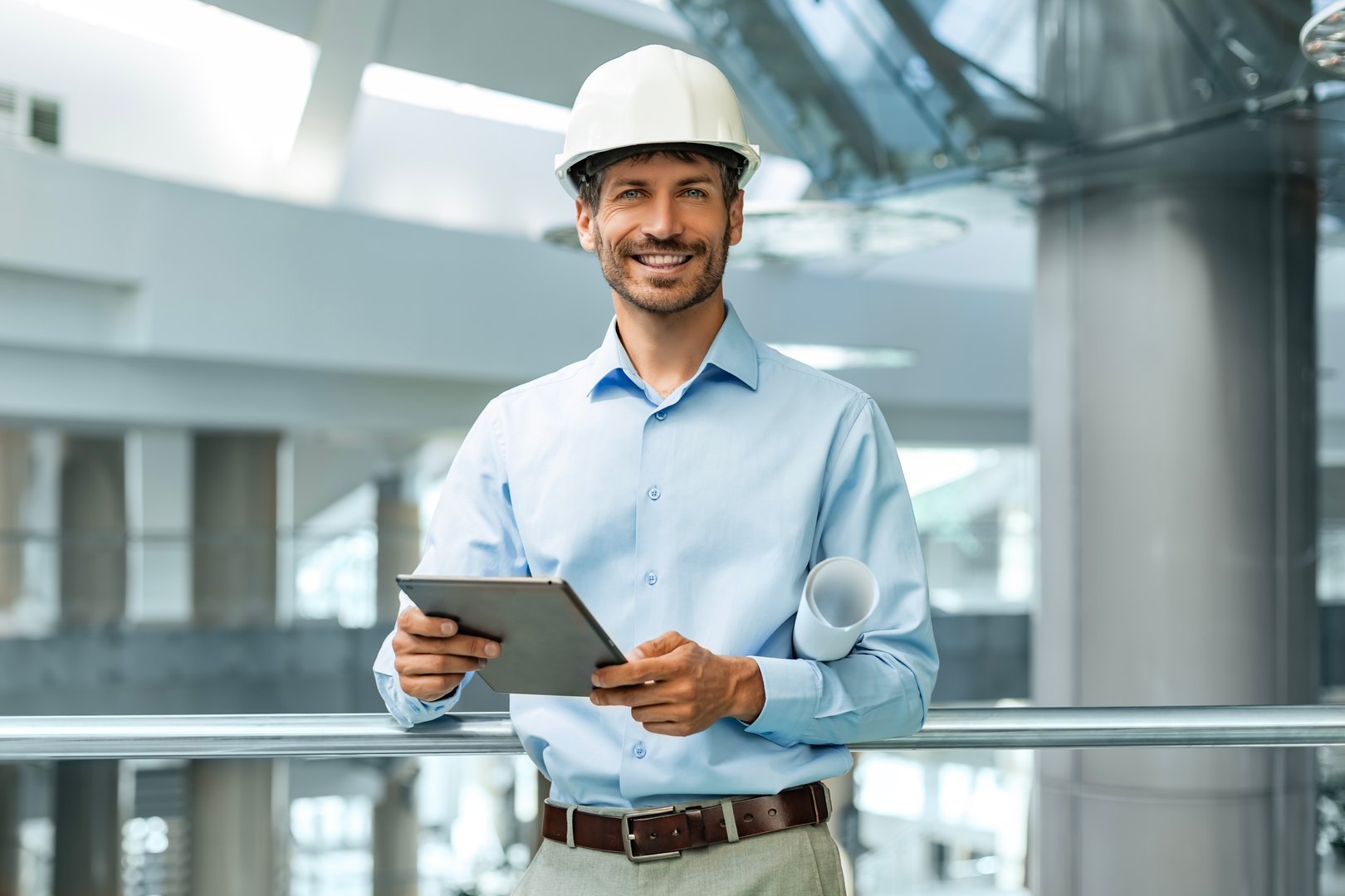 Handosme man with worker's helmet holding digital tablet while looking at camera in building under construction.
