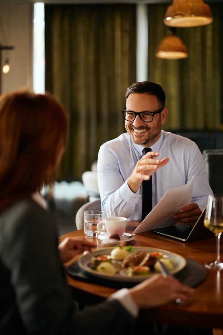 Happy entrepreneur talking to female coworker while having business lunch in restaurant.