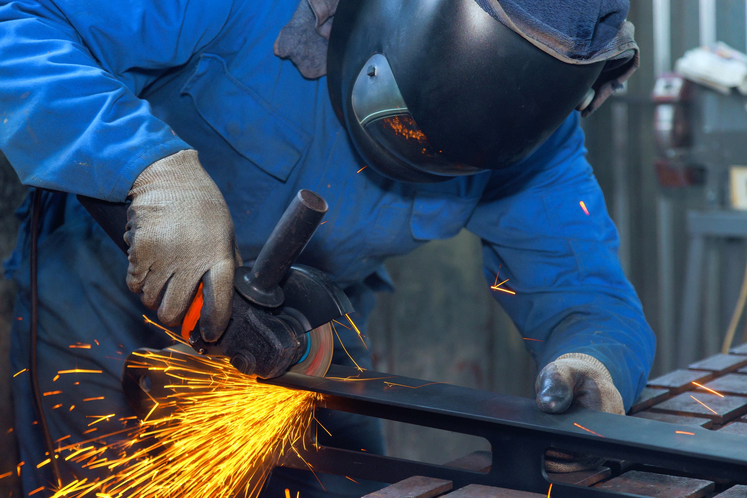 Worker in safety gear grinds metal, creating sparks, showcasing craftsmanship at metal workshop.