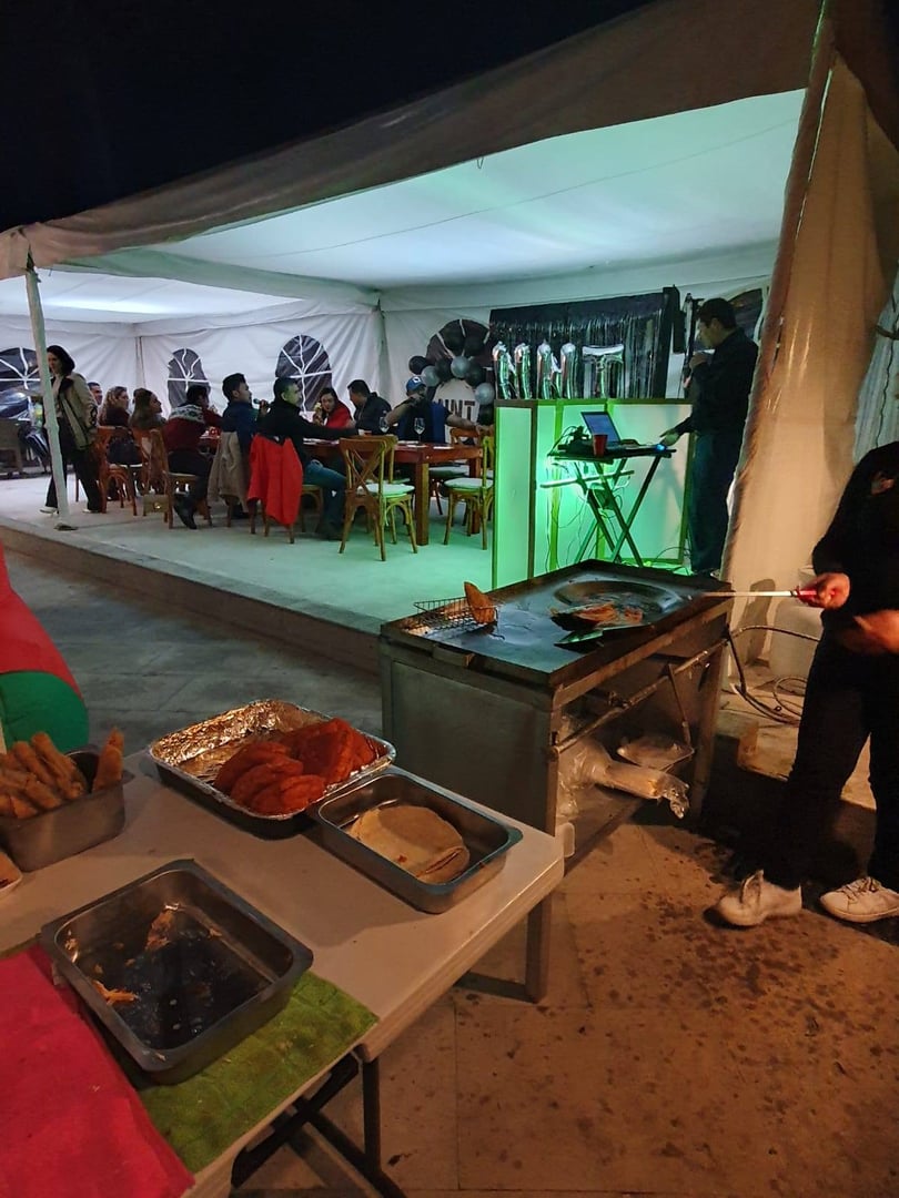 Outdoor event with food stand in foreground; people seated under a white tent with party lighting and decor.