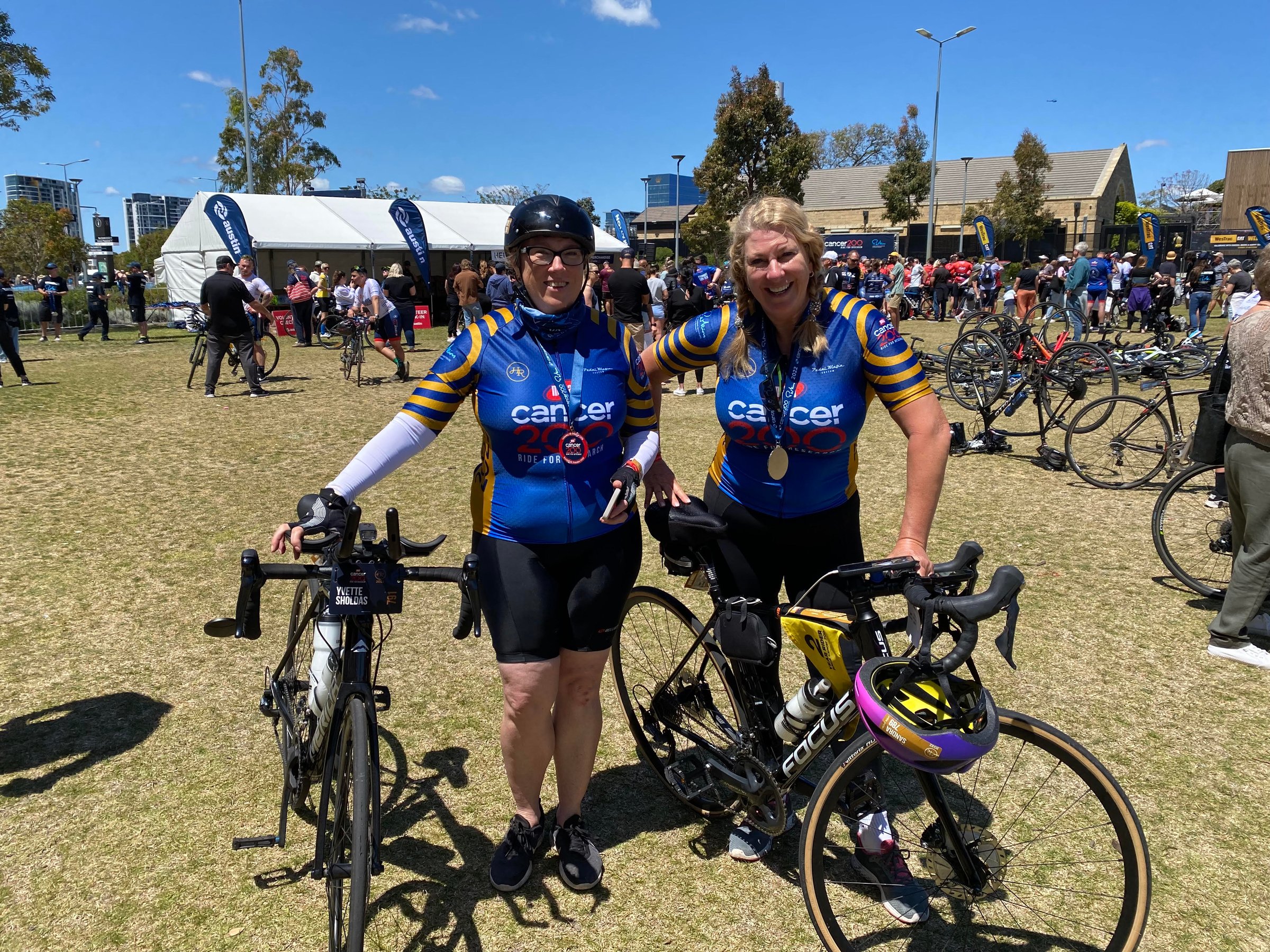 Two cyclists in matching jerseys pose with bikes at an outdoor cycling event; tents and participants are in the background.