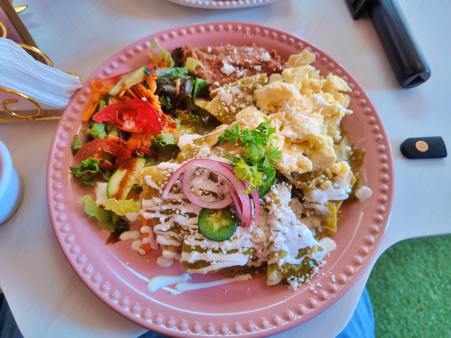 A vibrant and mouthwatering medium-shot photograph of a pink plate of Mexican green chilaquiles. The plate is piled high with crispy corn tortilla chips smothered in spicy green salsa, topped with scrambled eggs, crema, crumbled cotija cheese, slices of red onion, jalapeño pepper, and fresh cilantro. It's accompanied by a side of refried beans and a small fresh salad of lettuce, tomato, and cucumber. The scene captures an authentic and delicious homemade breakfast or brunch, with a focus on the texture, color, and freshness of the ingredients.