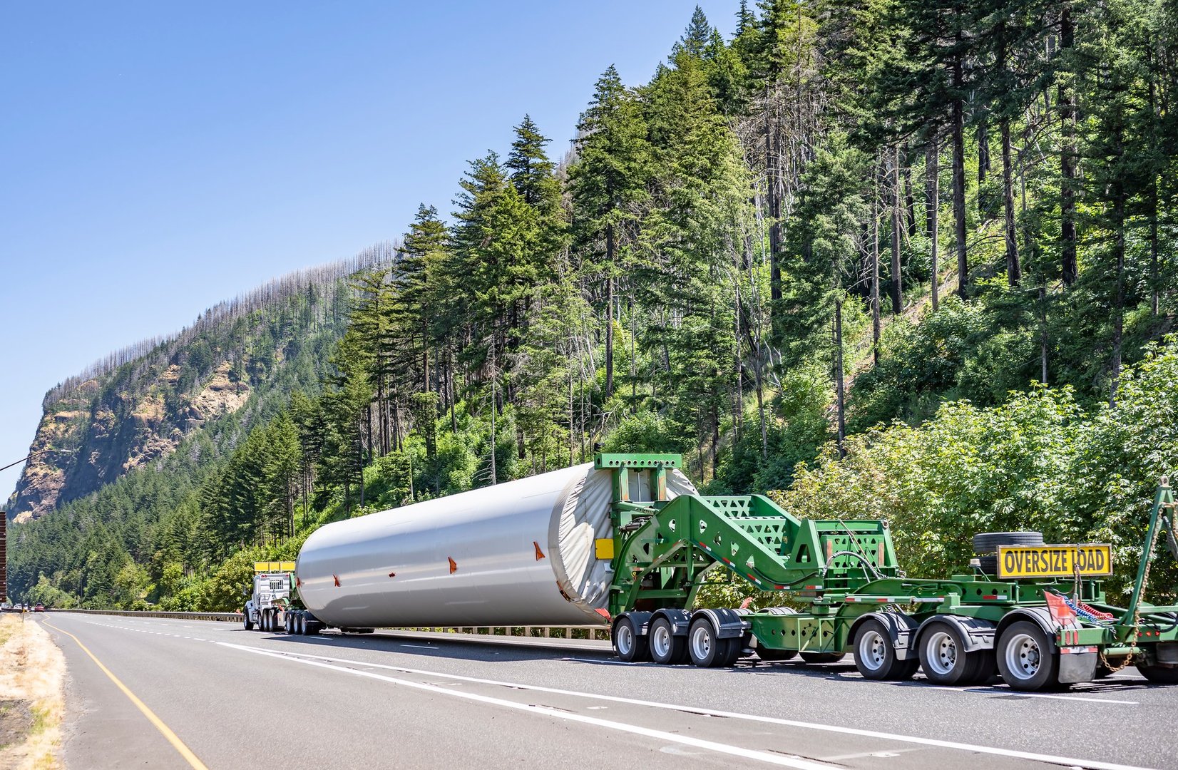 Heavy duty big rig semi truck transporting part of long oversized windmill electric generator pole on the specialized semi trailer with additional trolleys and oversize load sign running on the road