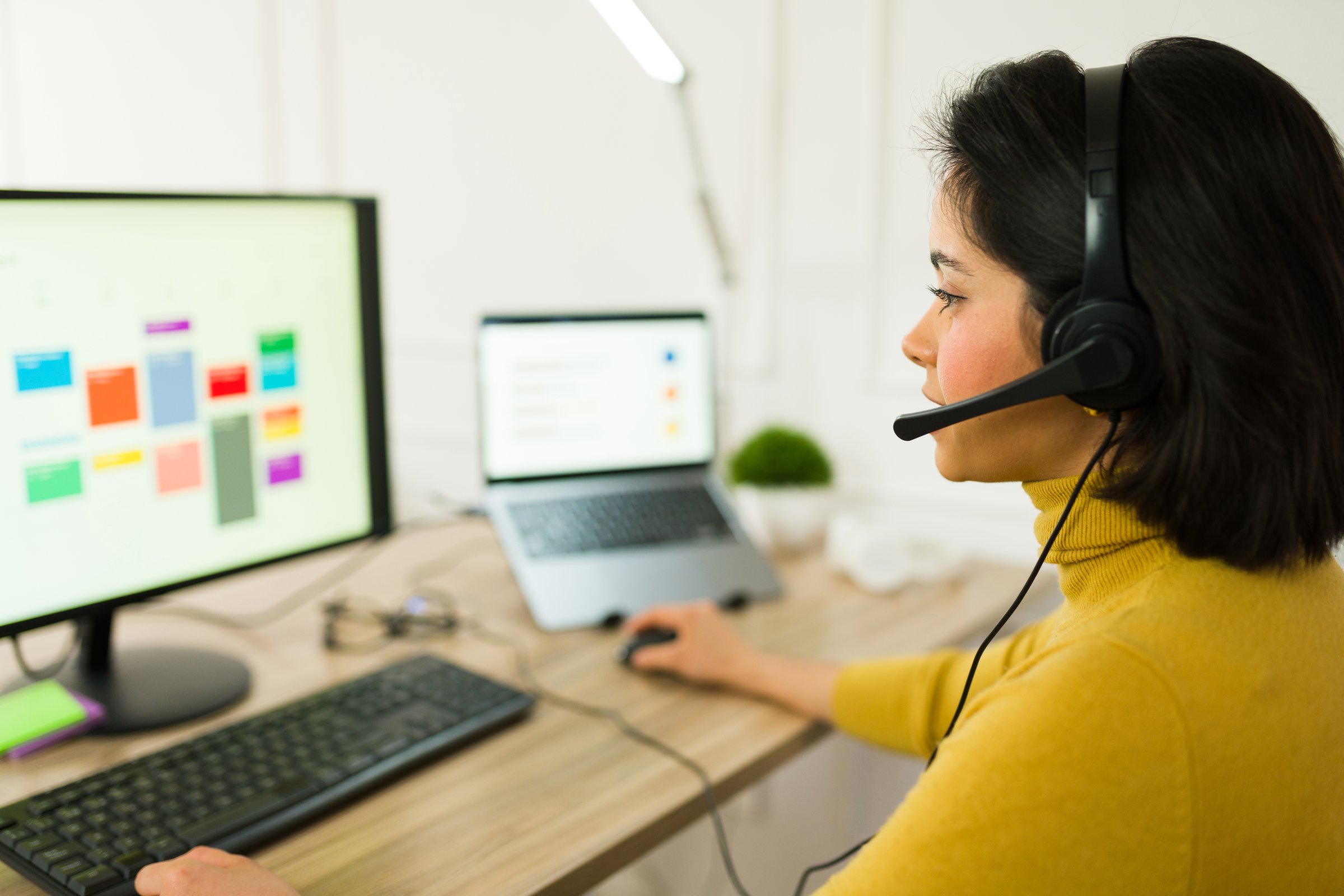Latin freelancer wearing wireless headset typing on laptop, managing remote project and providing customer support from home workspace