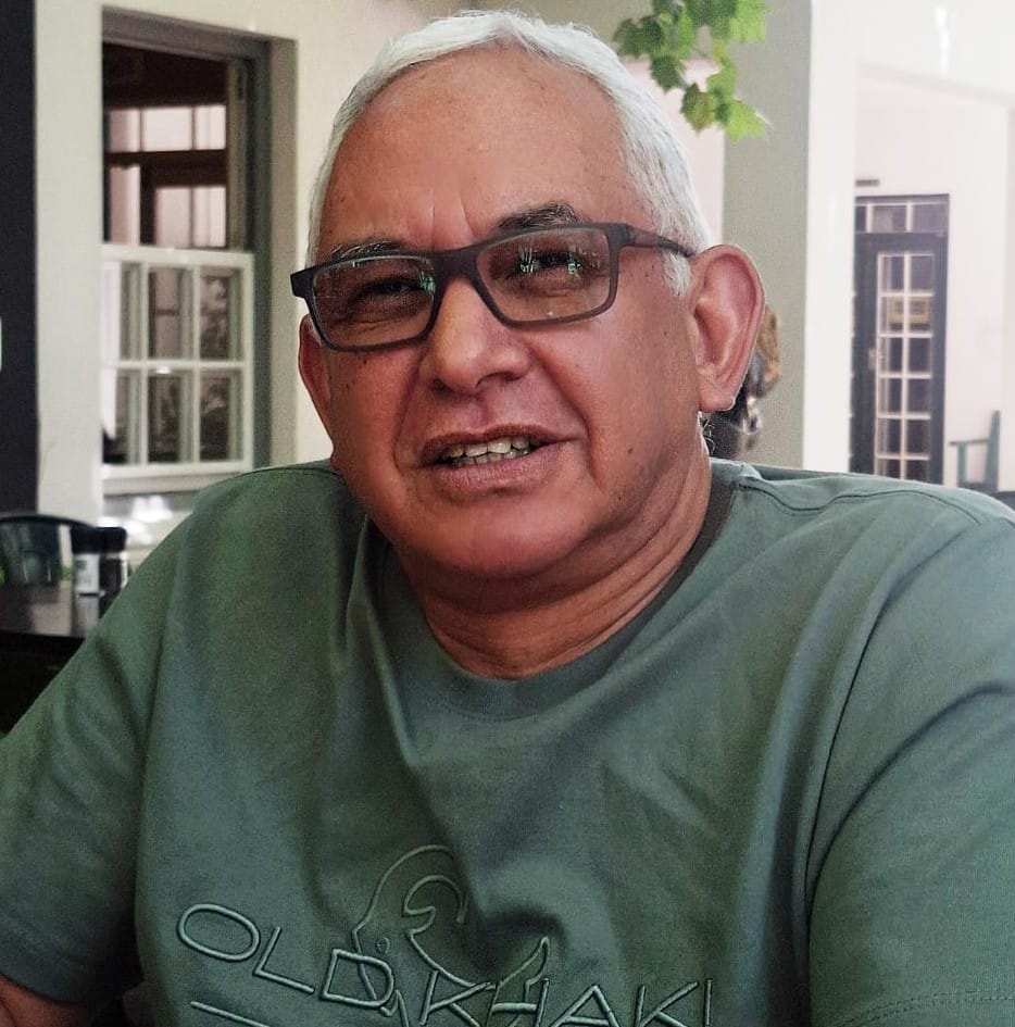 Smiling man with white hair and glasses wearing a green T-shirt sitting indoors.