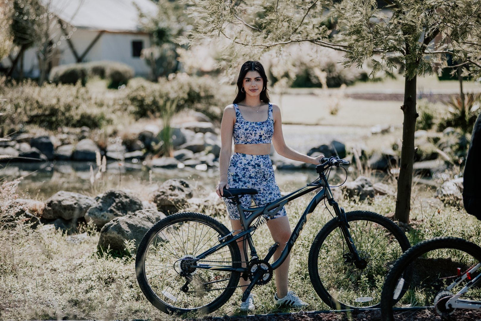 A woman in a blue floral outfit stands next to a bicycle outdoors, surrounded by trees and a rocky area in the background.